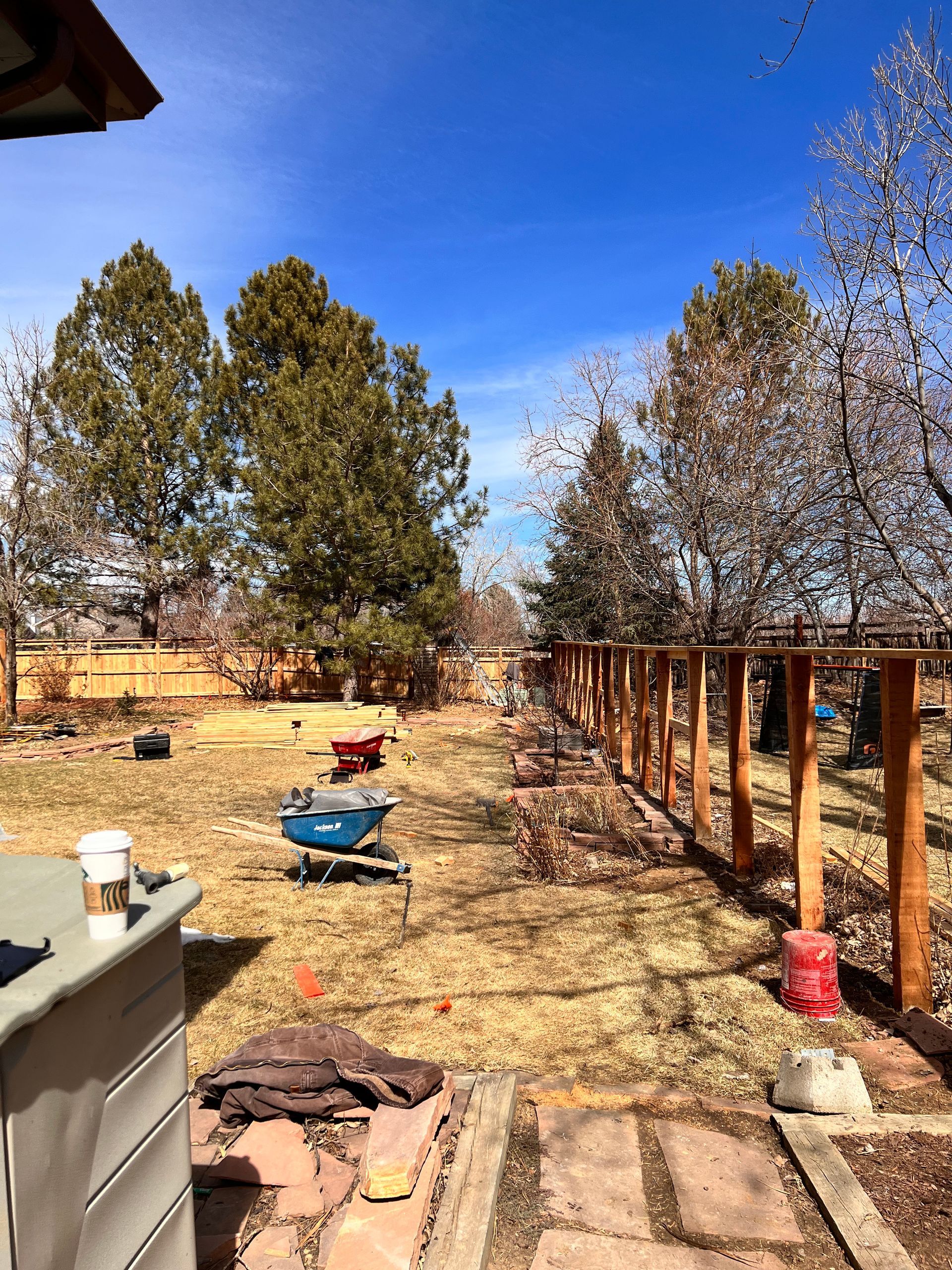 Backyard with a partially built wooden fence. Green trees, blue sky, and construction tools are visible.