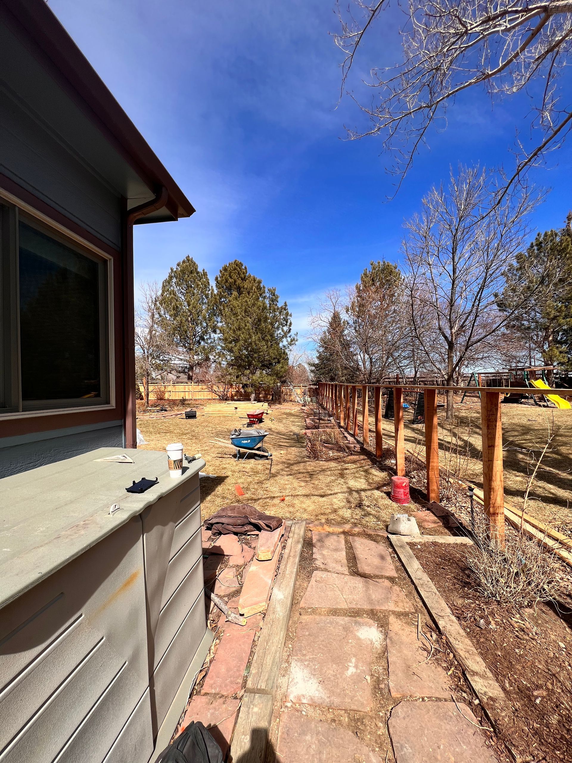Backyard view with fence under construction, brown grass, blue sky, and a house on the left.