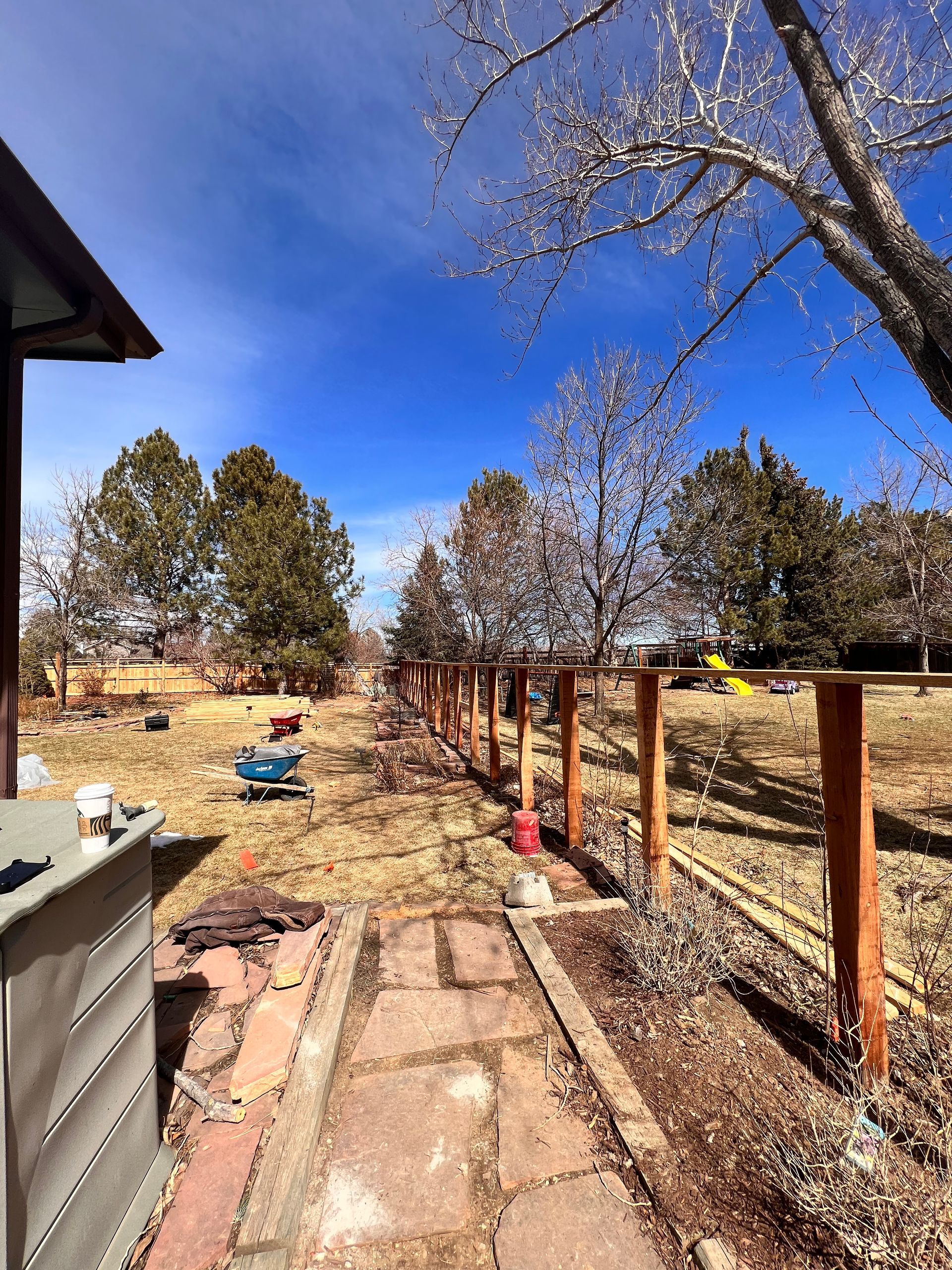 Backyard with a brick path, fence posts, trees, and bright blue sky.
