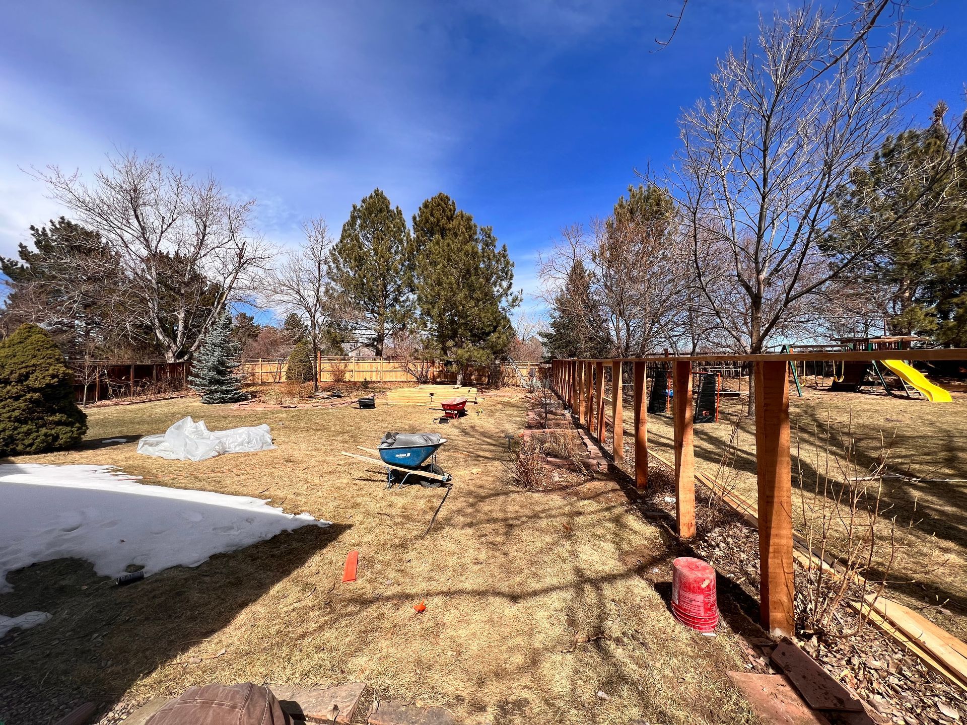 Backyard with a partially built wooden fence; blue sky; snow patches; wheelbarrow, tools, and trees.