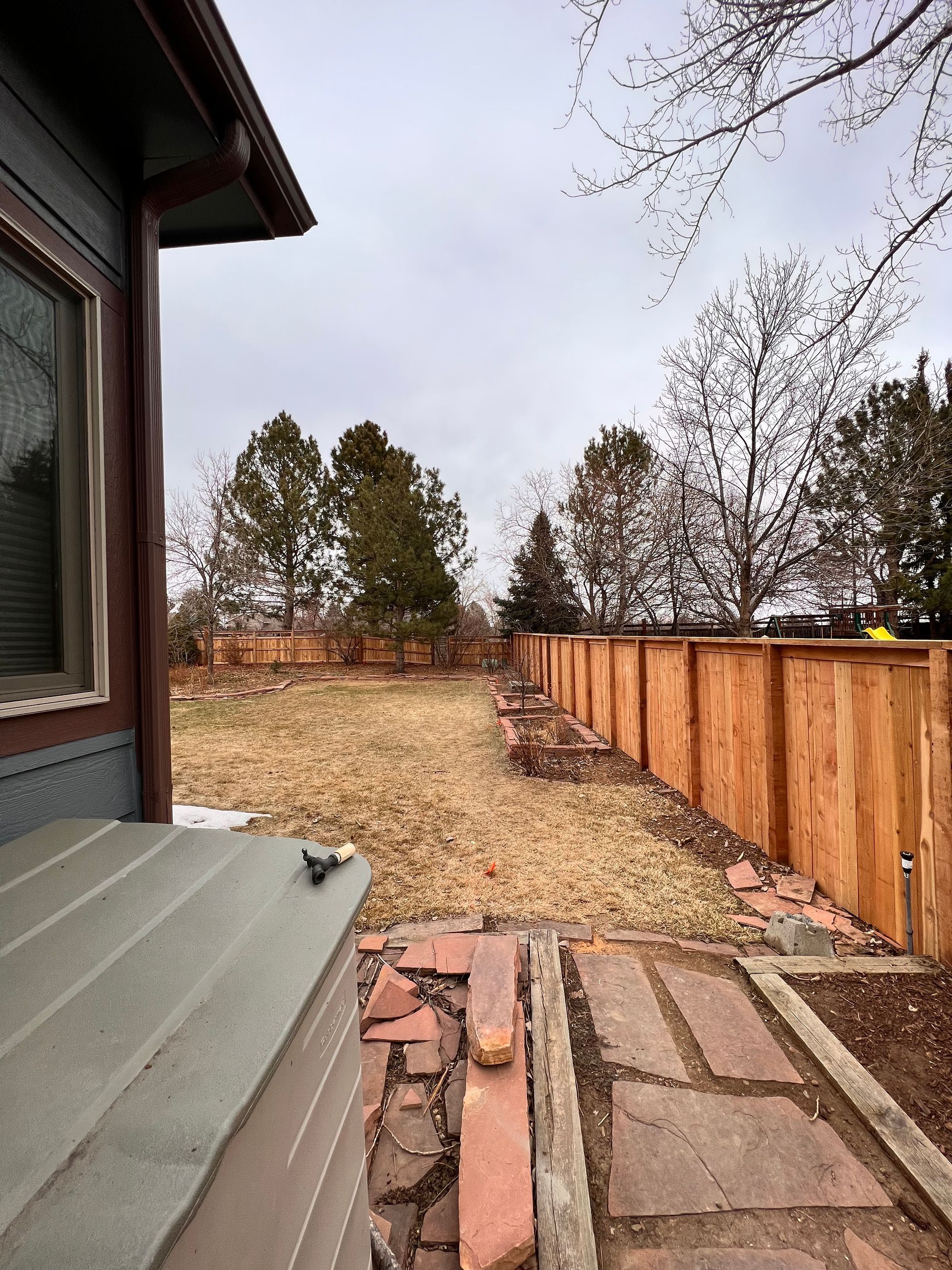 Backyard view: brown wooden fence, dry grass, brick path, trees, overcast sky.