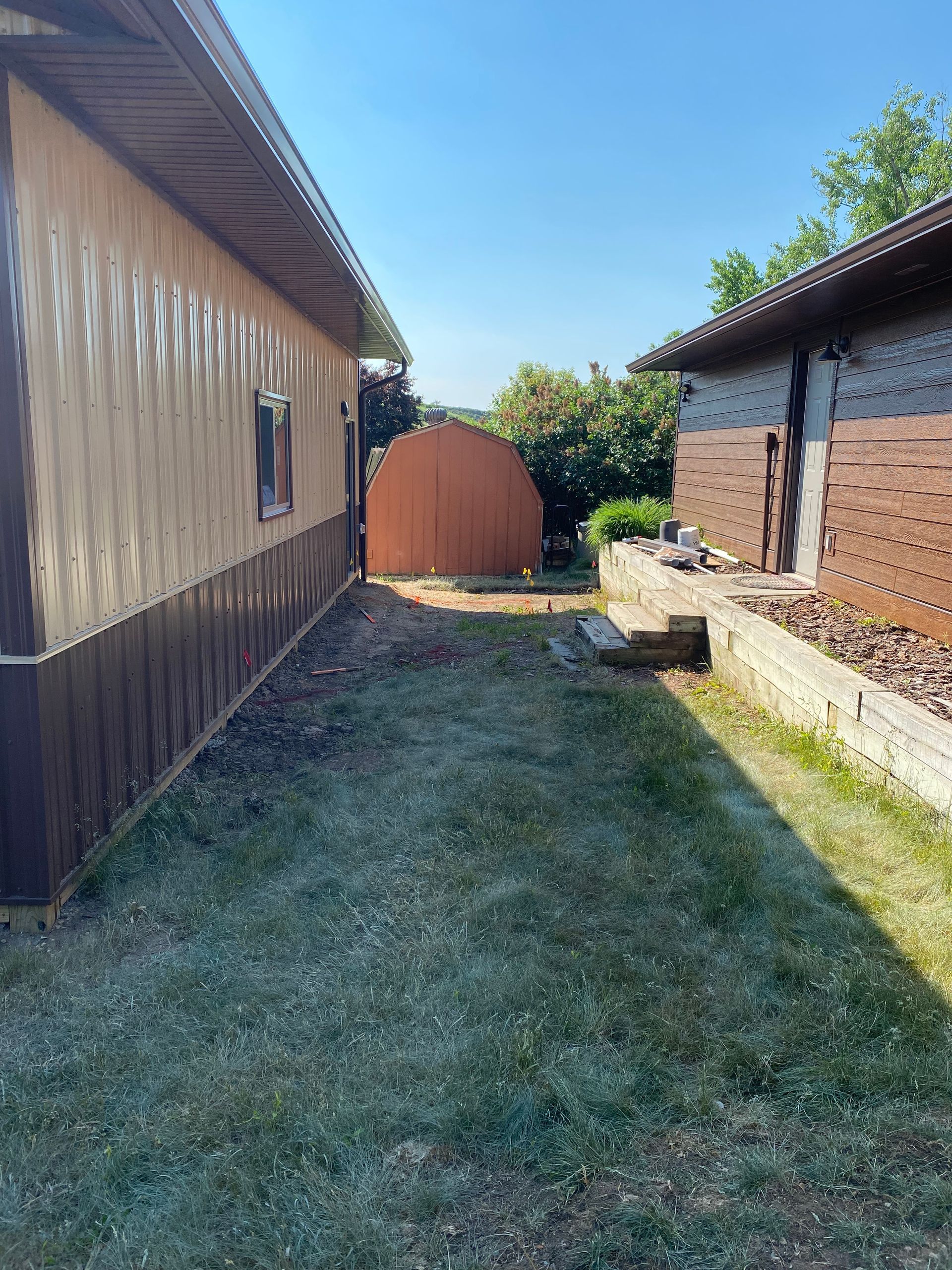Narrow, grassy yard between two buildings, one with tan siding, the other with brown siding. A brown dome structure is visible.