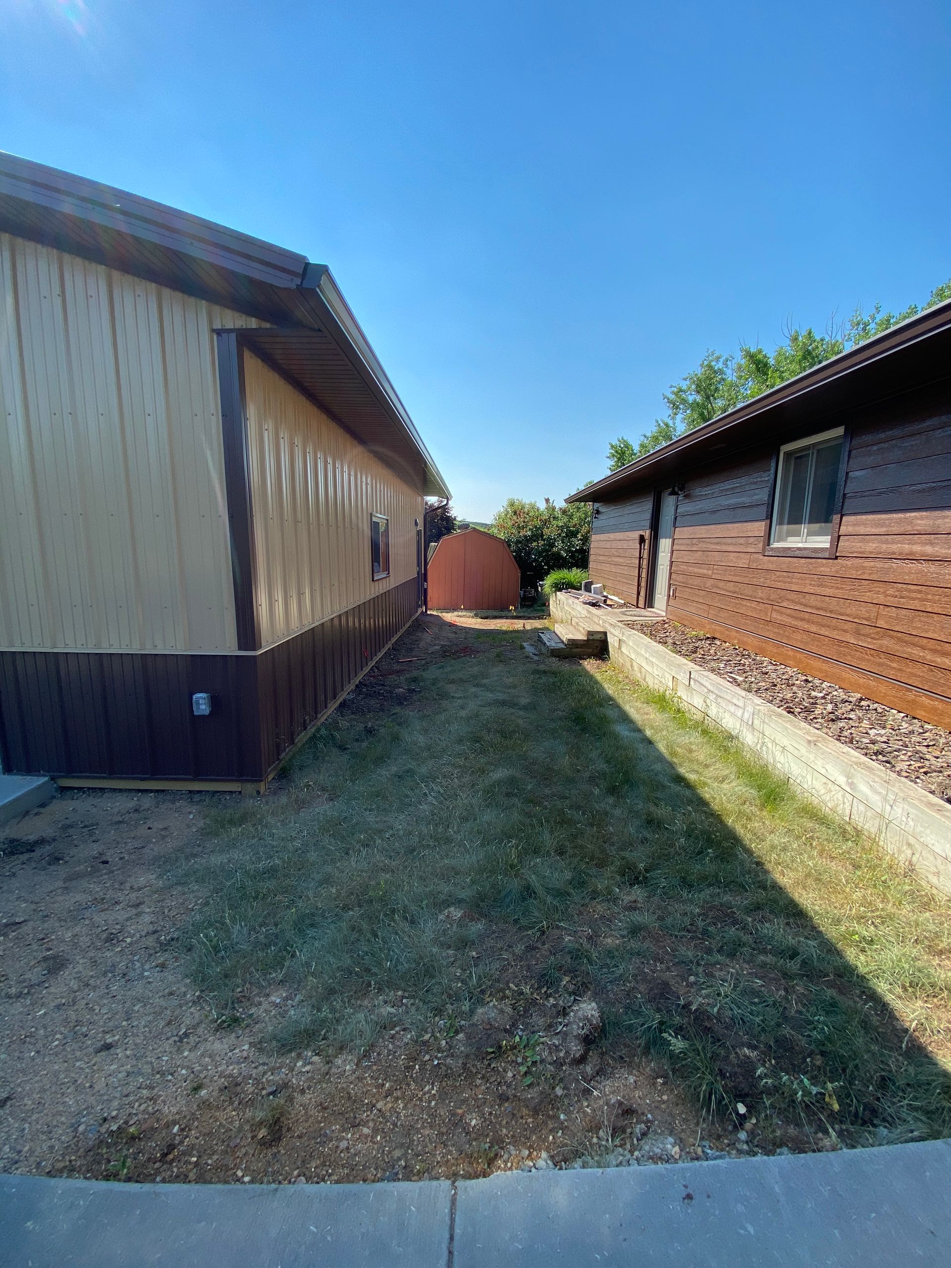 Narrow grassy path between two buildings, one with brown siding, the other with brick, under a clear blue sky.