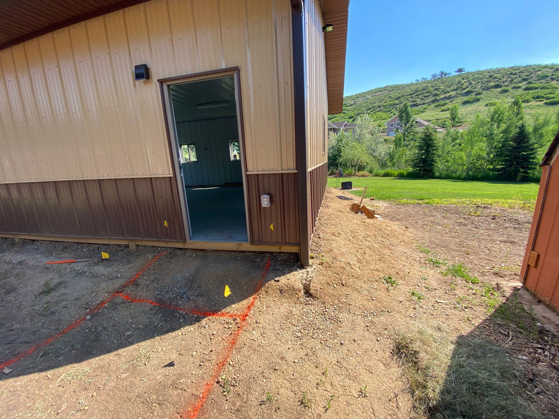 Tan and brown building with open doorway. Dirt ground with orange lines, green hillside in the distance.