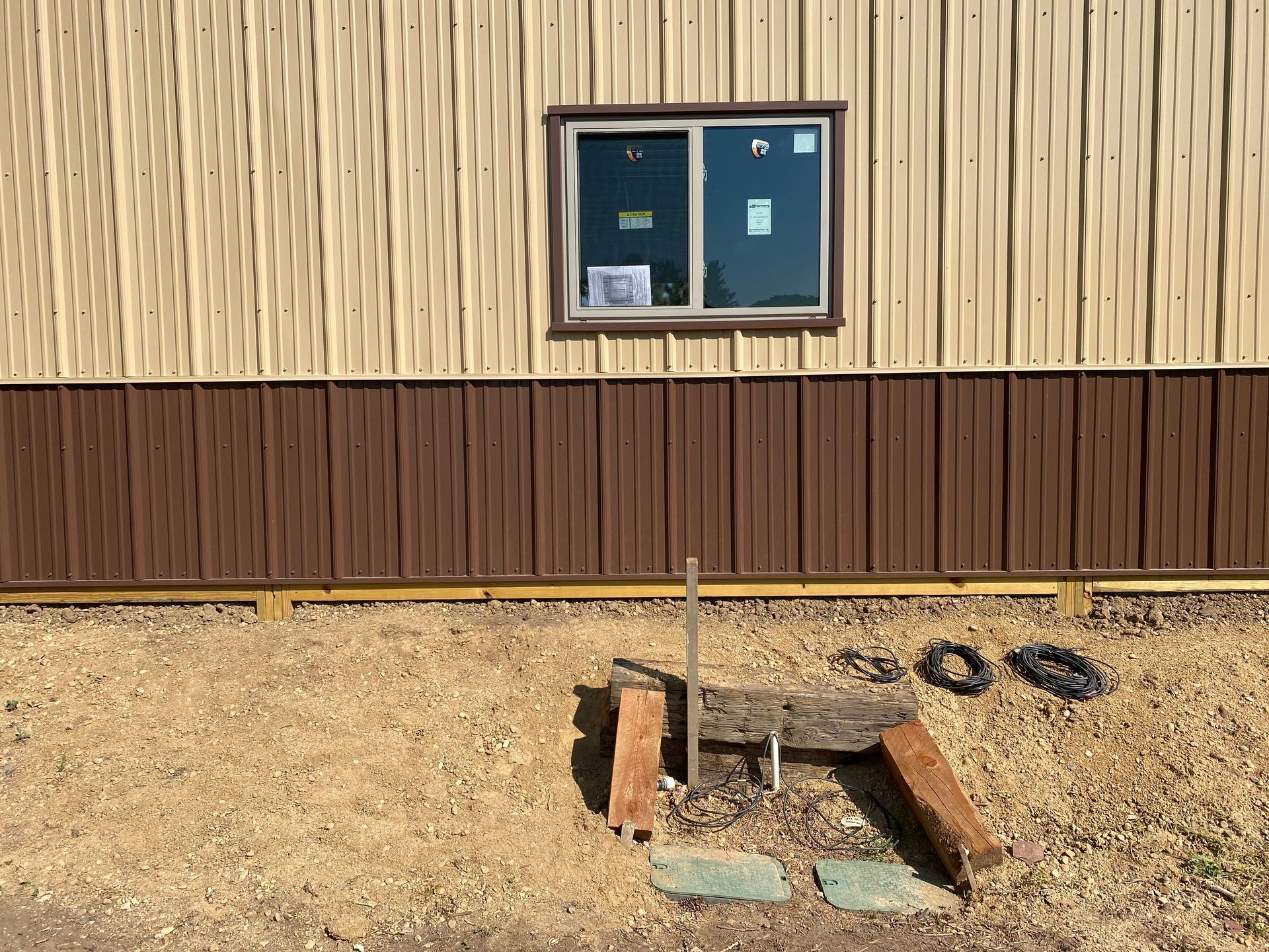 Tan and brown building with a window, brown trim, and a small wooden structure on a gravel ground.