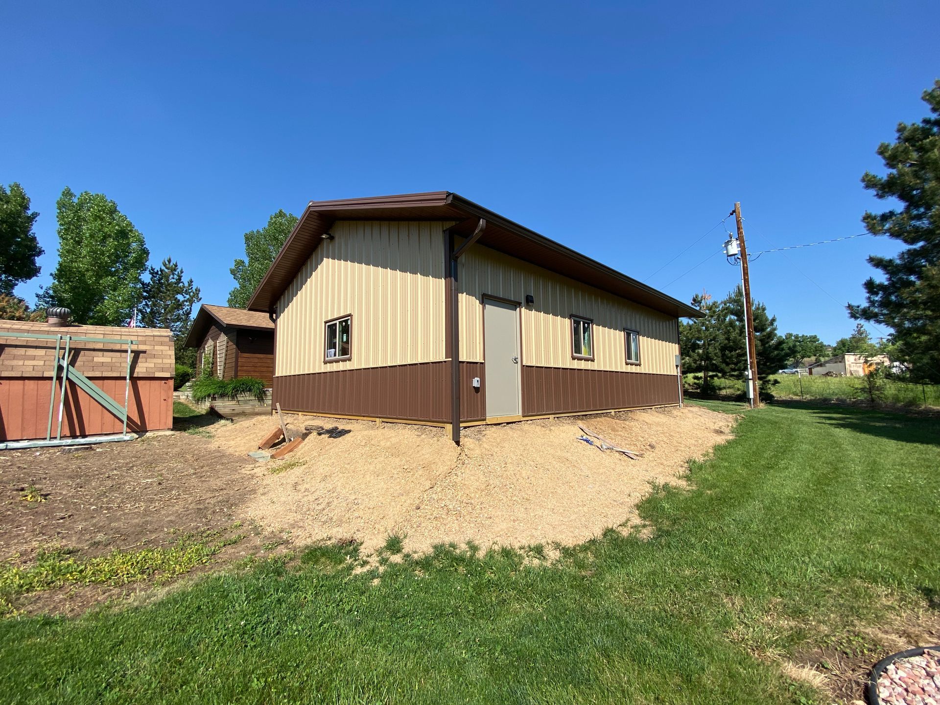Brown and tan metal building with door, small windows, and gravel base on a sunny day.