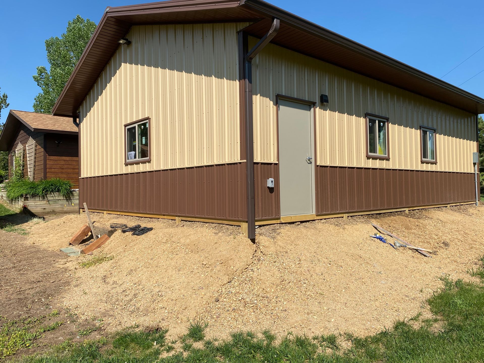 Tan and brown building with a gravel base, a door, and two windows. A smaller building is in the background.