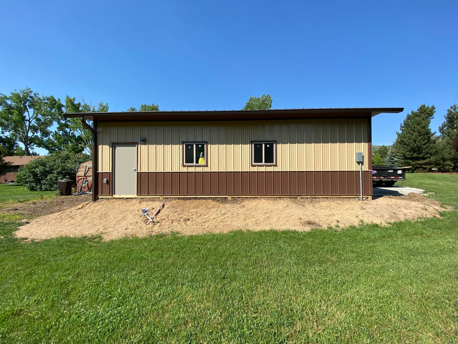 Building with brown siding, two windows, and a door, set in a grassy area with a blue sky.