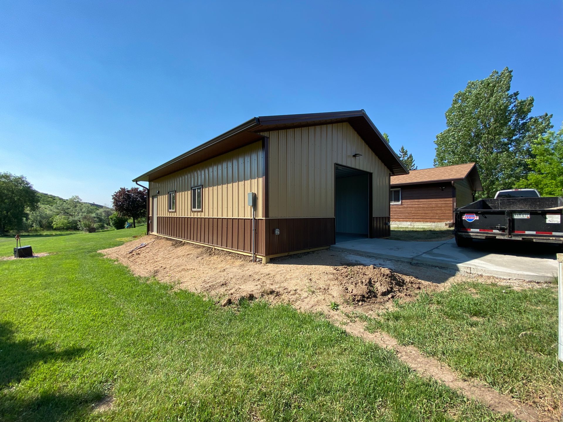 Tan and brown metal garage with open door on a grassy lot under a blue sky.