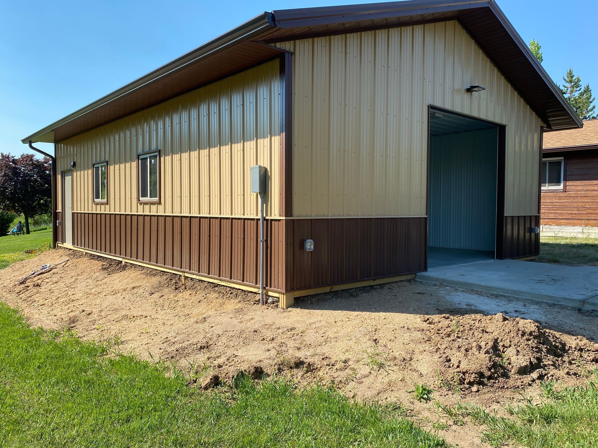 Tan and brown metal-sided garage with open bay door on concrete pad; dirt and grass in foreground.