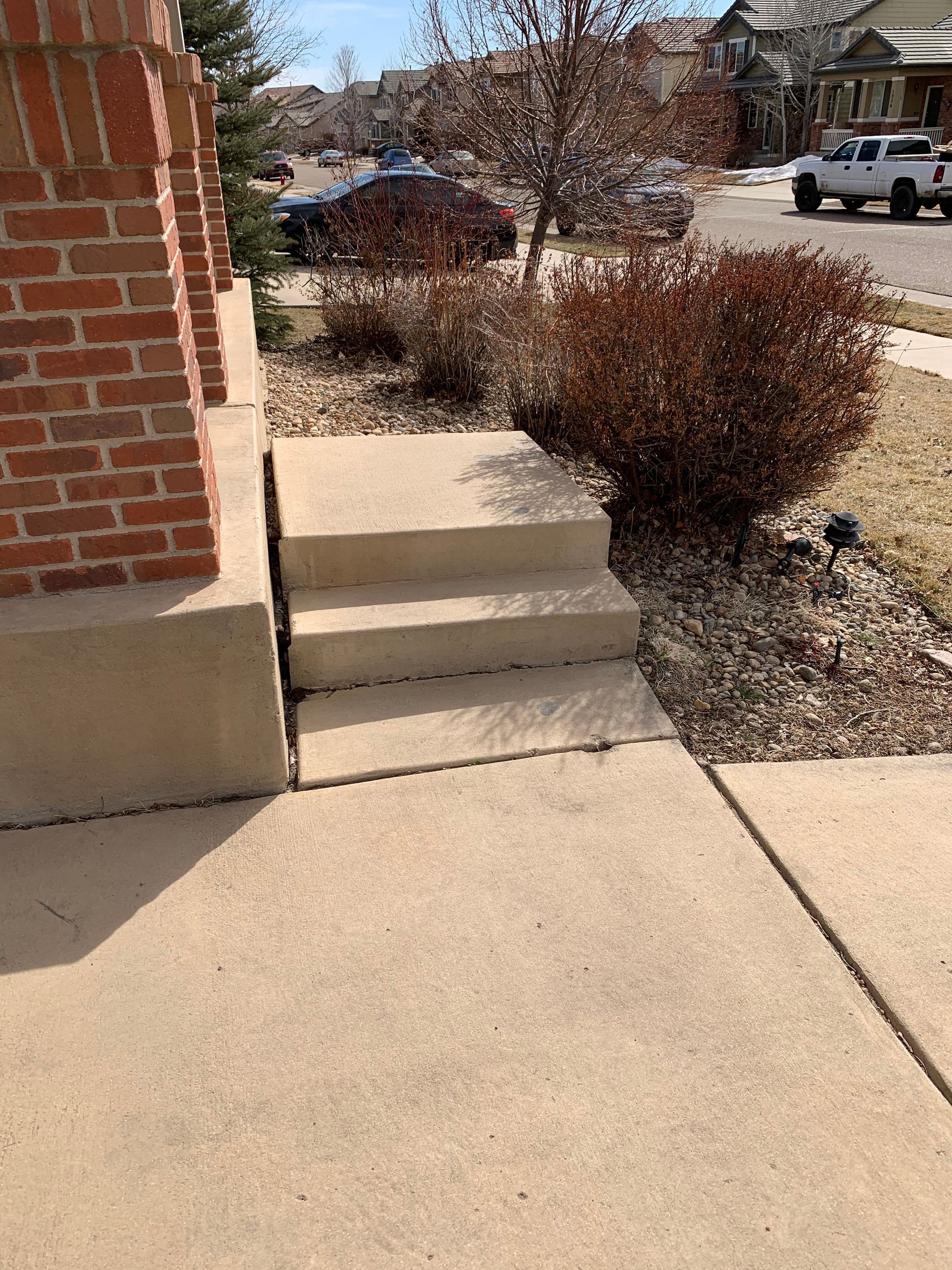 Concrete steps leading to a brick pillar porch, sidewalk in the foreground, bushes and street in the background.