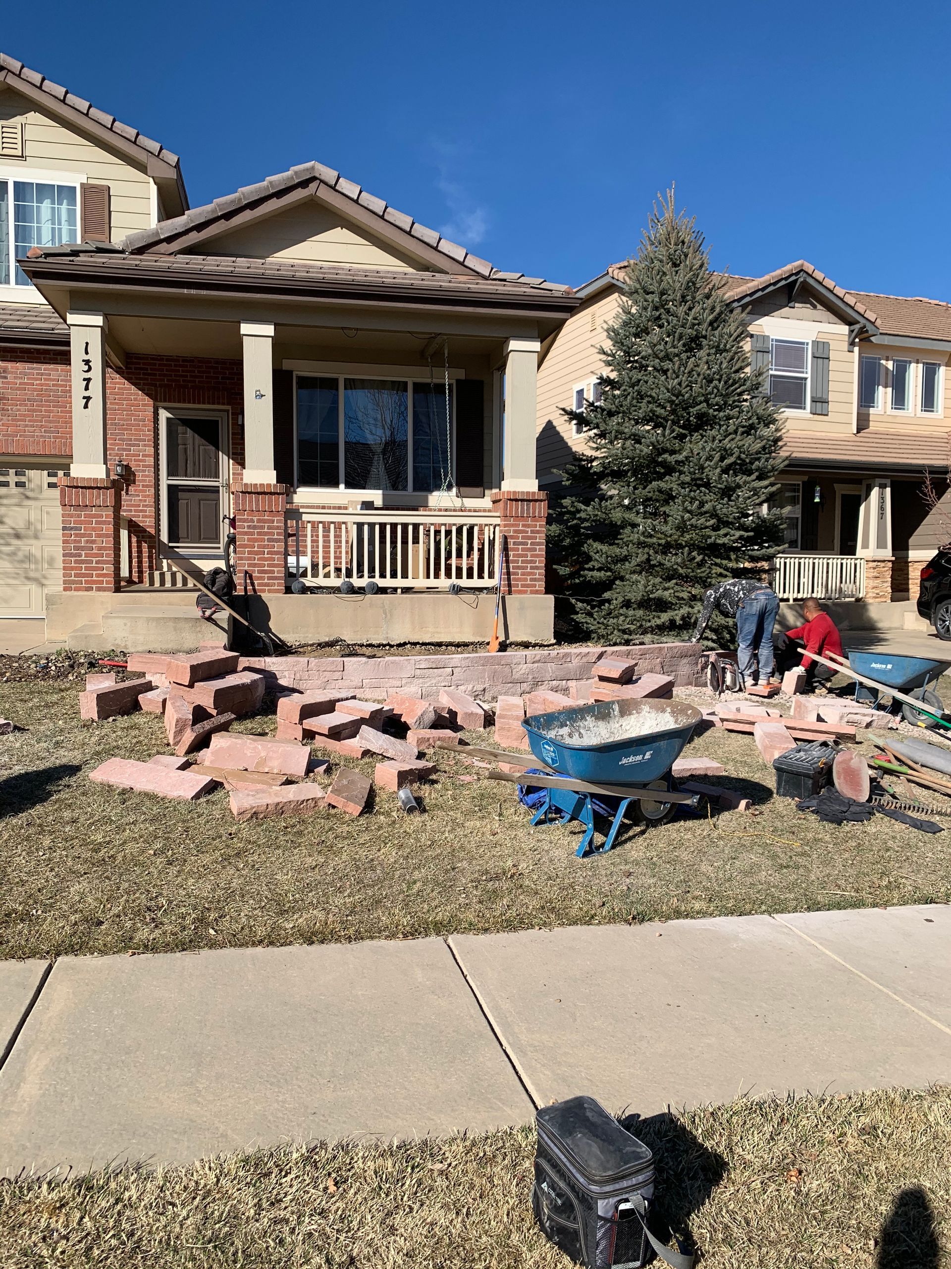Brick pathway construction in front of a house. Workers, tools, materials scattered on the lawn. Sunny day.