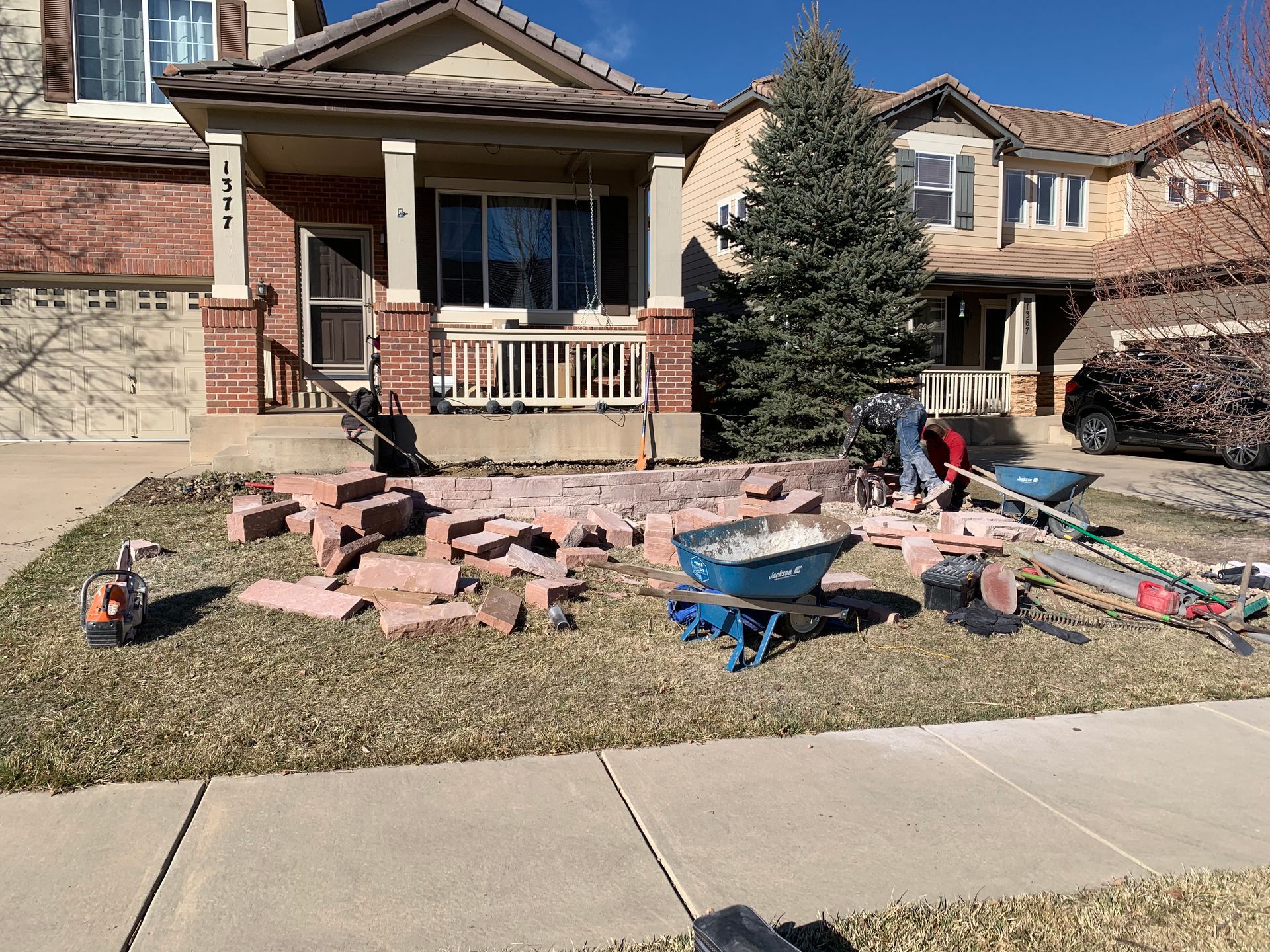 Workers rebuilding a brick front yard feature in front of a house, scattered brick and tools.