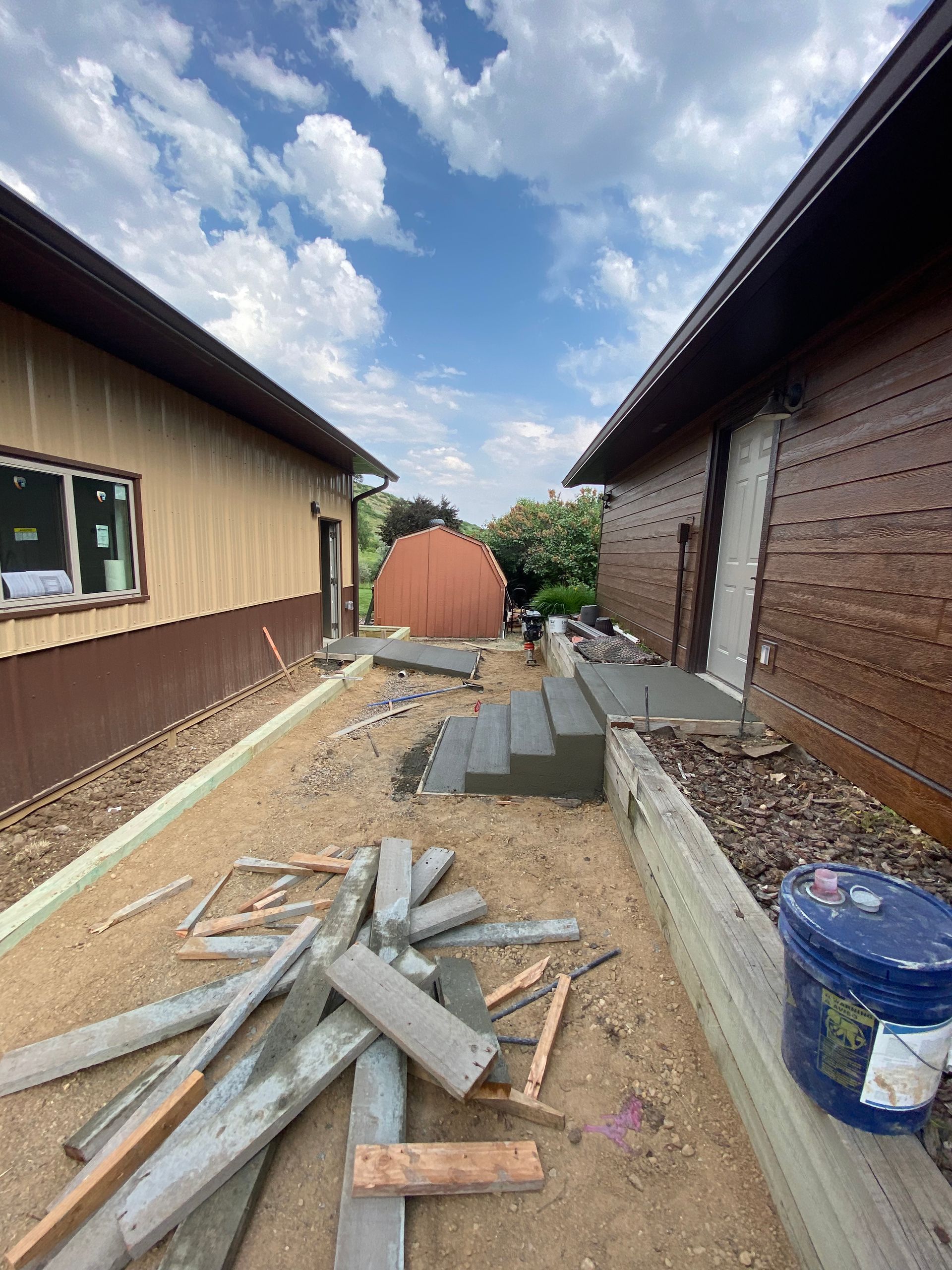 Pathway construction between two buildings; gravel, concrete steps, and blue bucket.