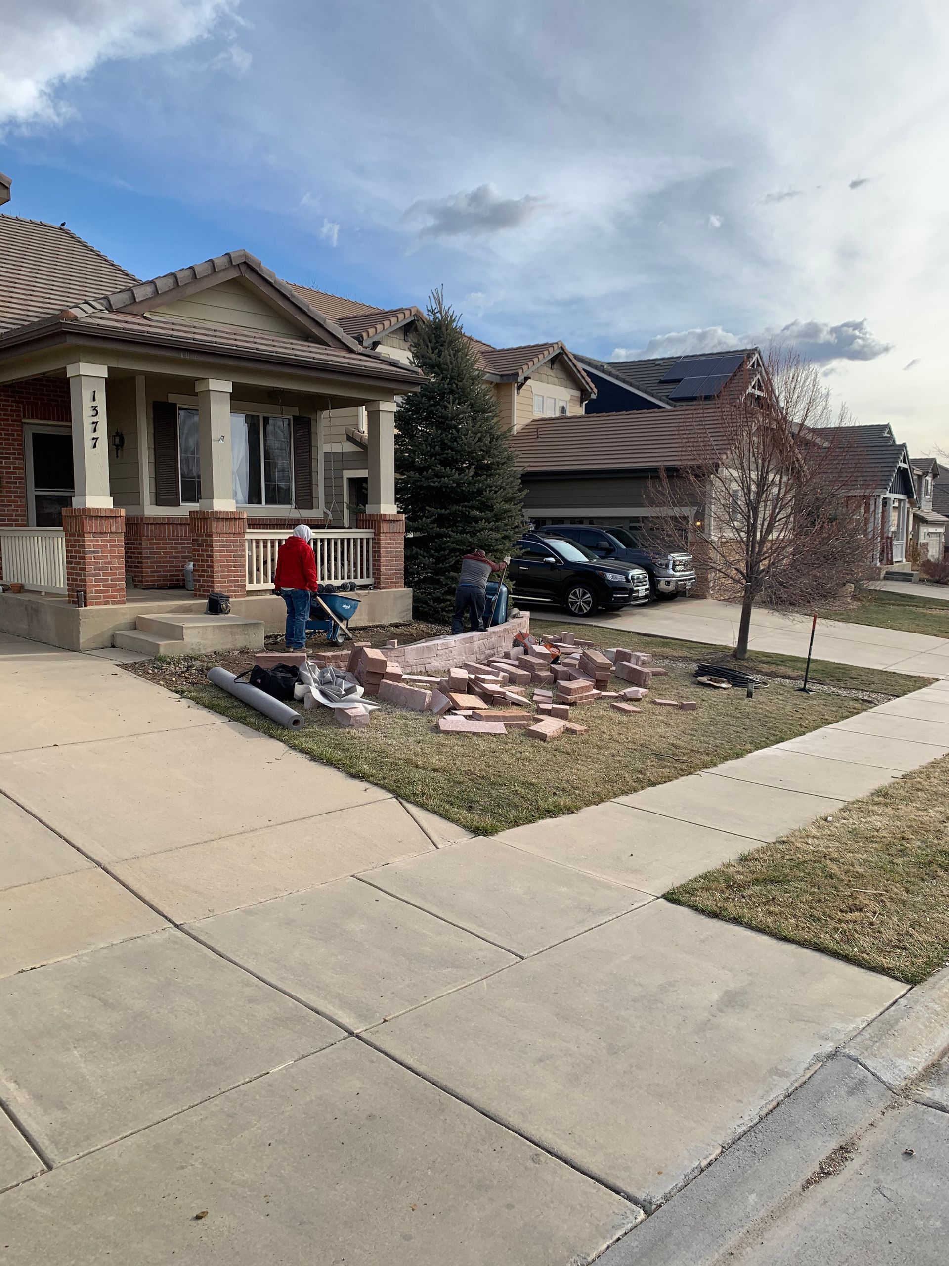 People working on a house's front yard, bricks in foreground, residential neighborhood.