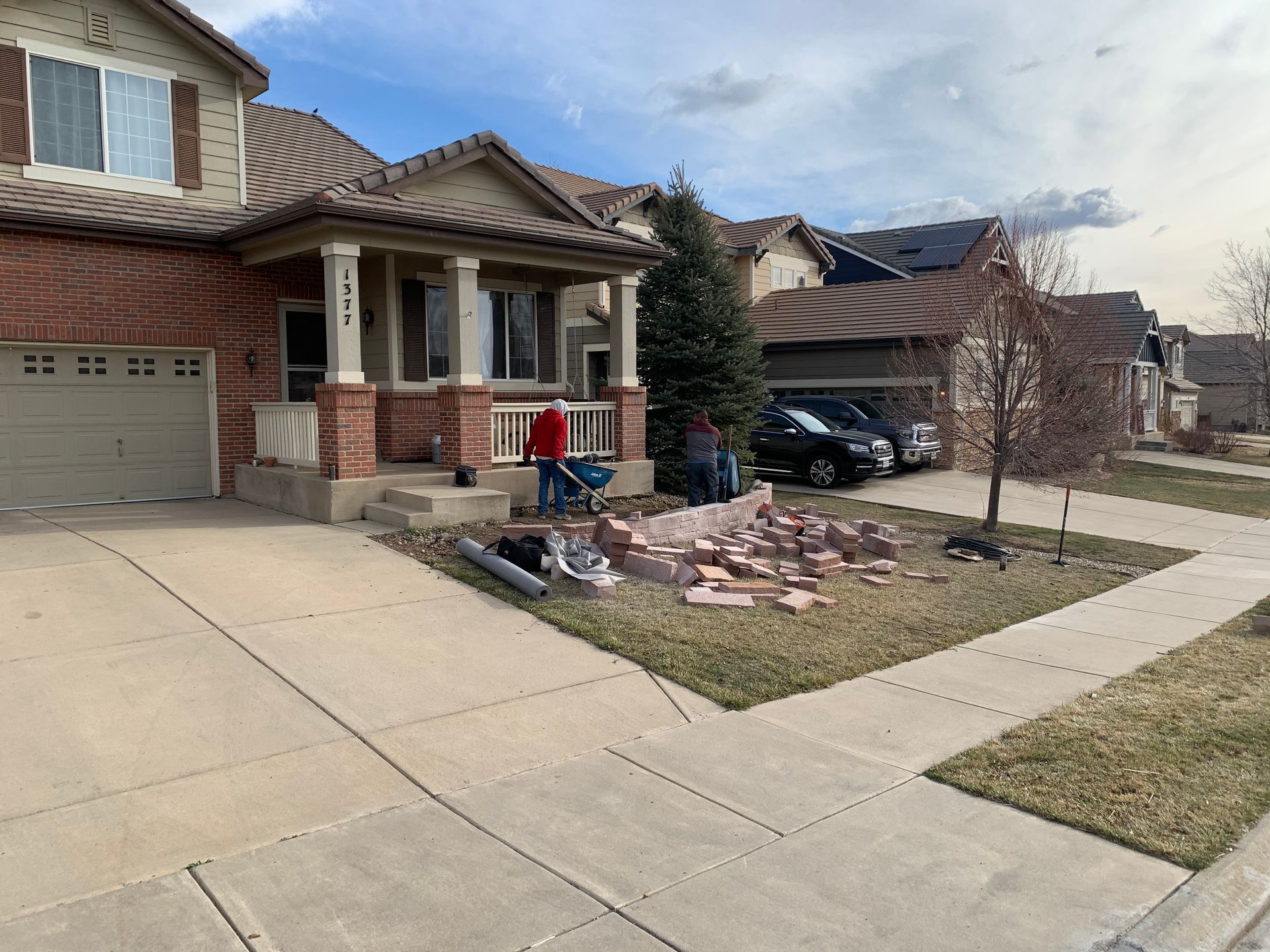 Two people working on a brick retaining wall in front of a house on a sunny day.