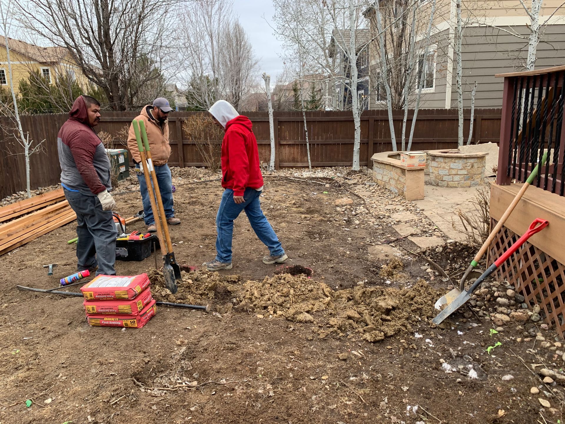 Three people digging in a backyard with tools and building materials; preparing for construction.