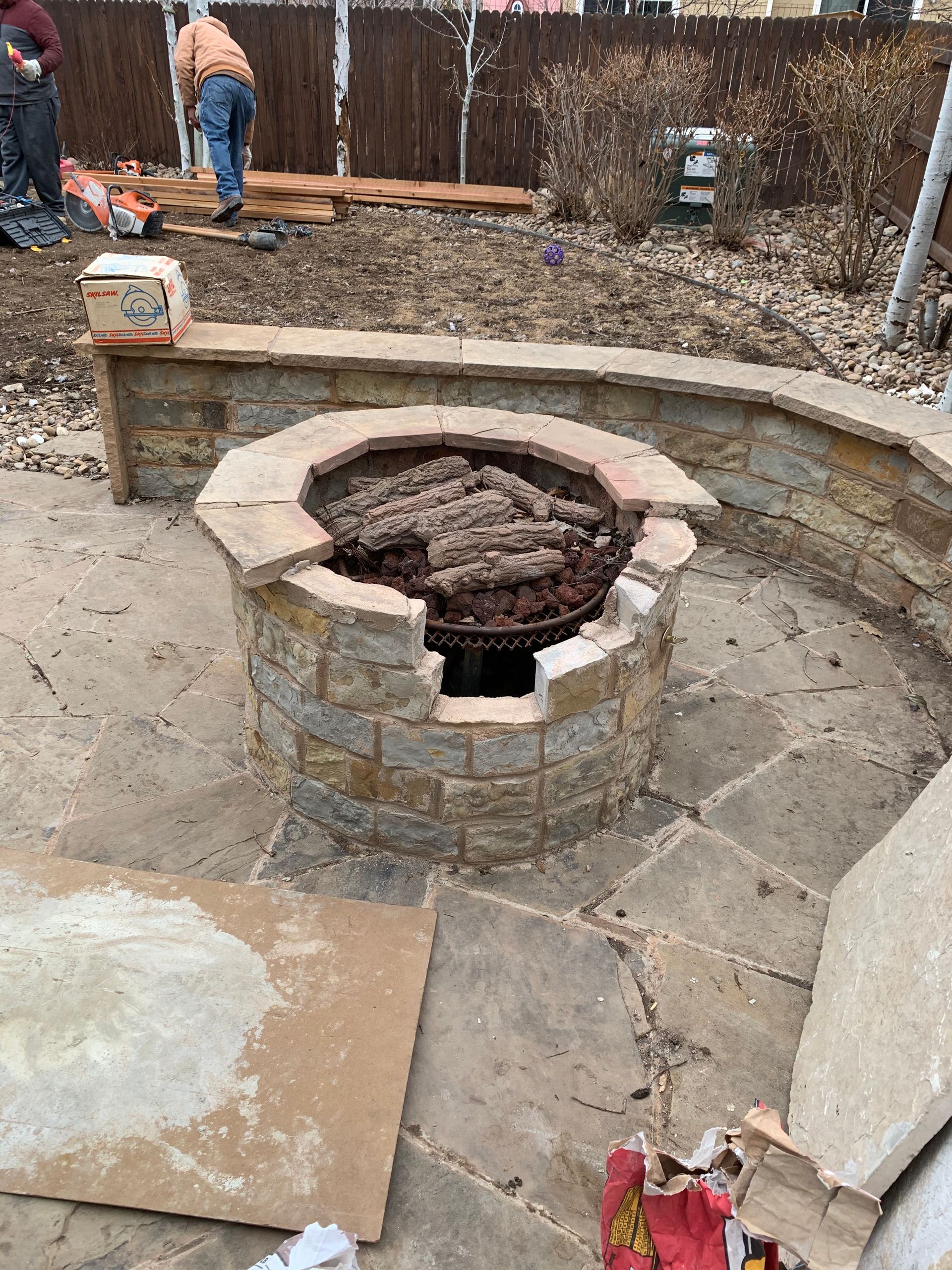 Stone fire pit construction in progress, with pavers and brick walls, surrounded by landscaping.