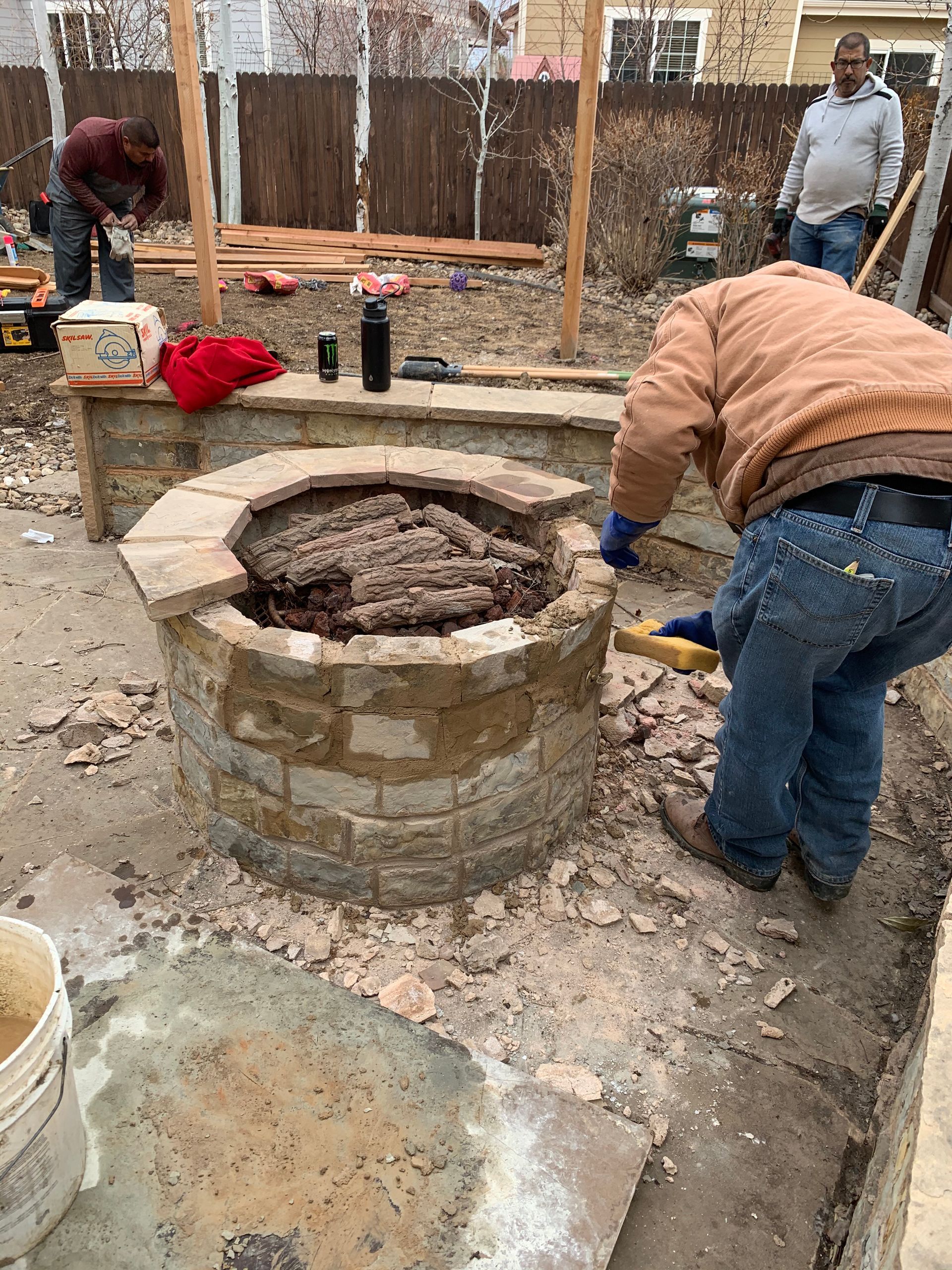 Men building a brick fire pit in a backyard, using tools and materials.
