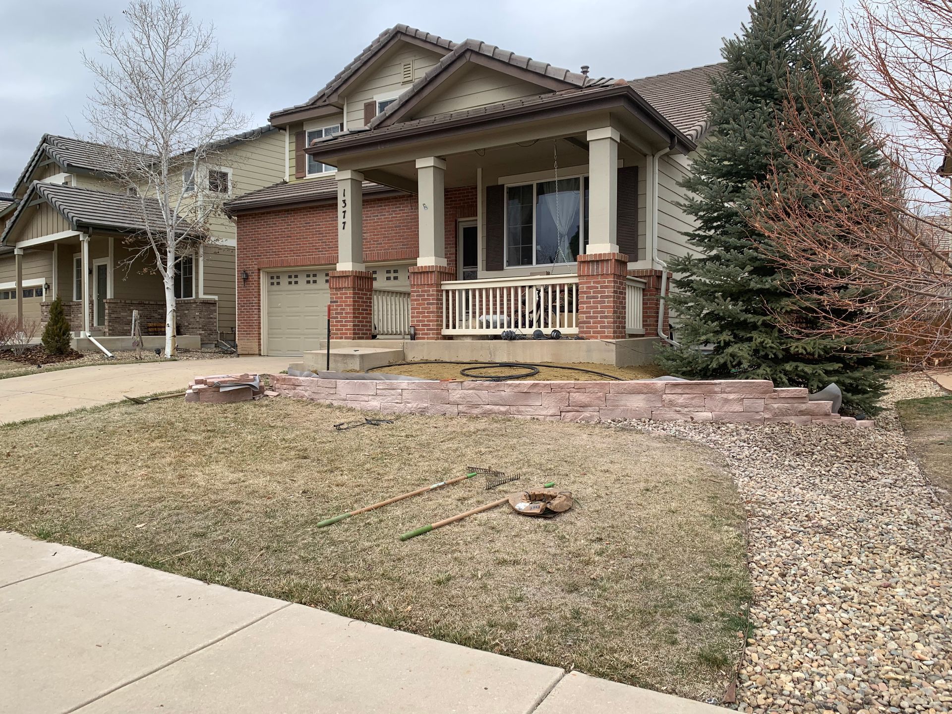 A two-story house with a brick facade and a porch. A retaining wall and dead grass in front.