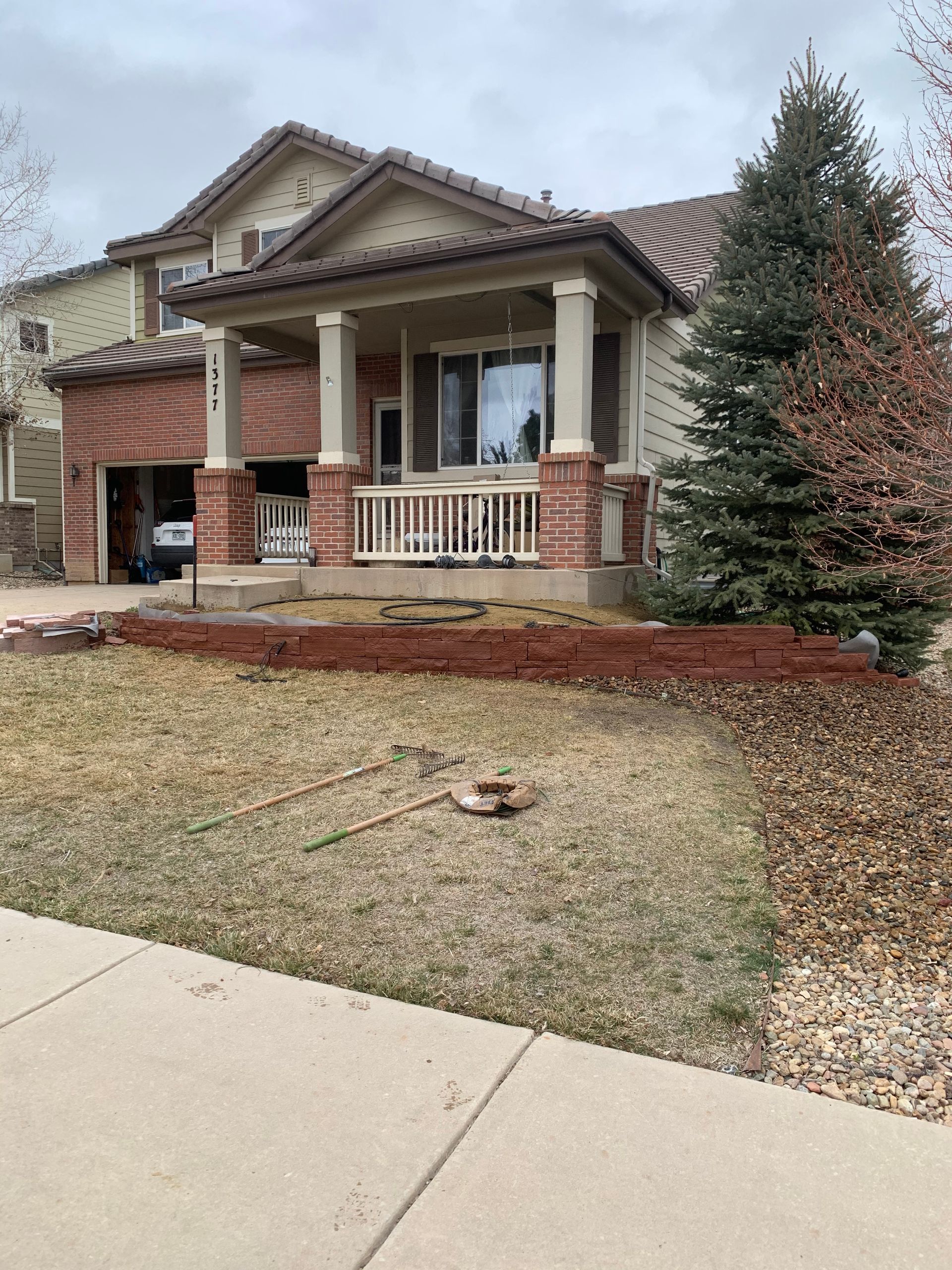 Two-story house with a brick facade and porch, brown mulch, and green lawn. Overcast sky.