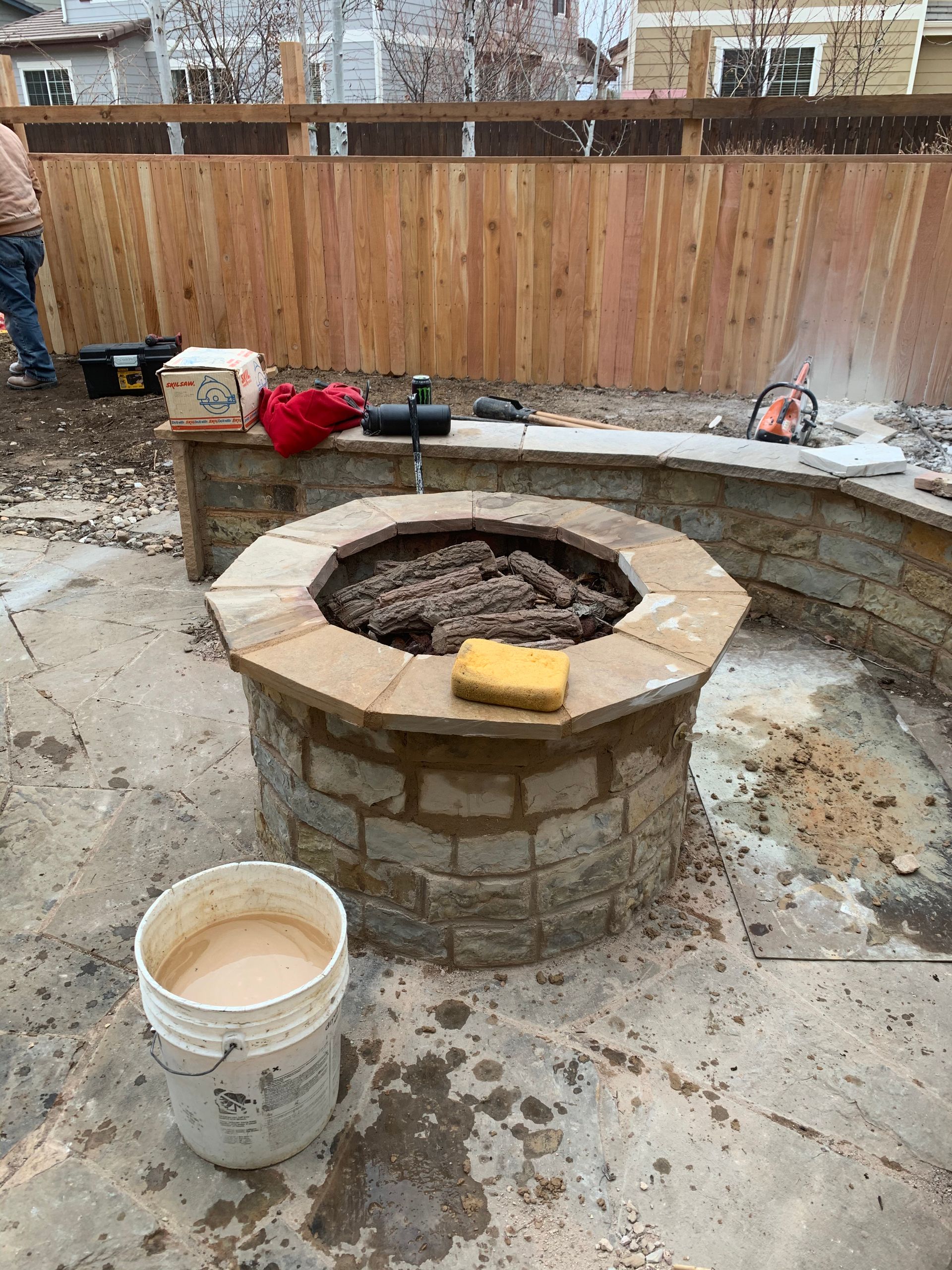 Stone fire pit under construction in a yard, with a fence in the background and a bucket of mortar in the foreground.
