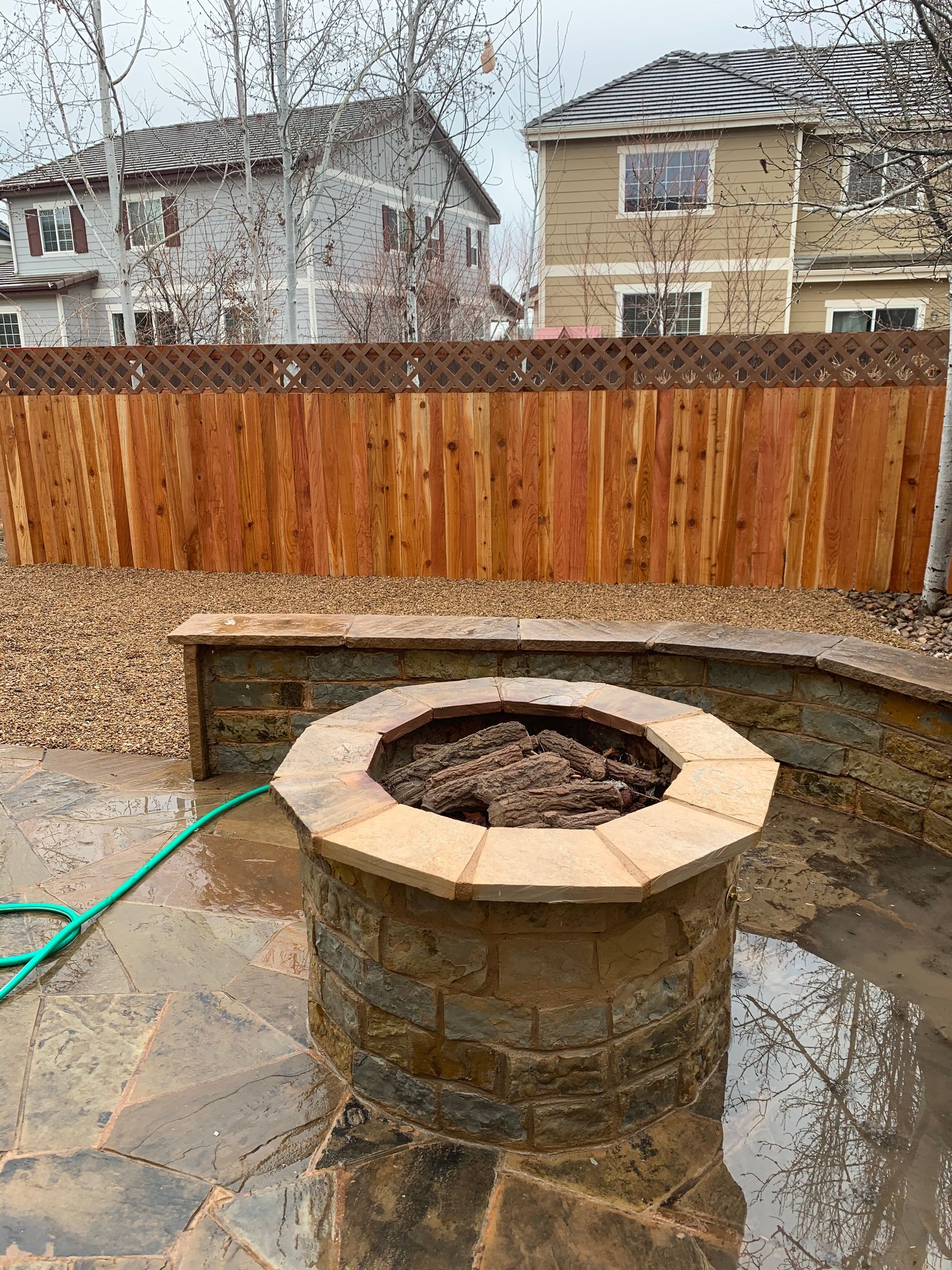 Stone fire pit on a patio with a wooden fence and houses in the background.