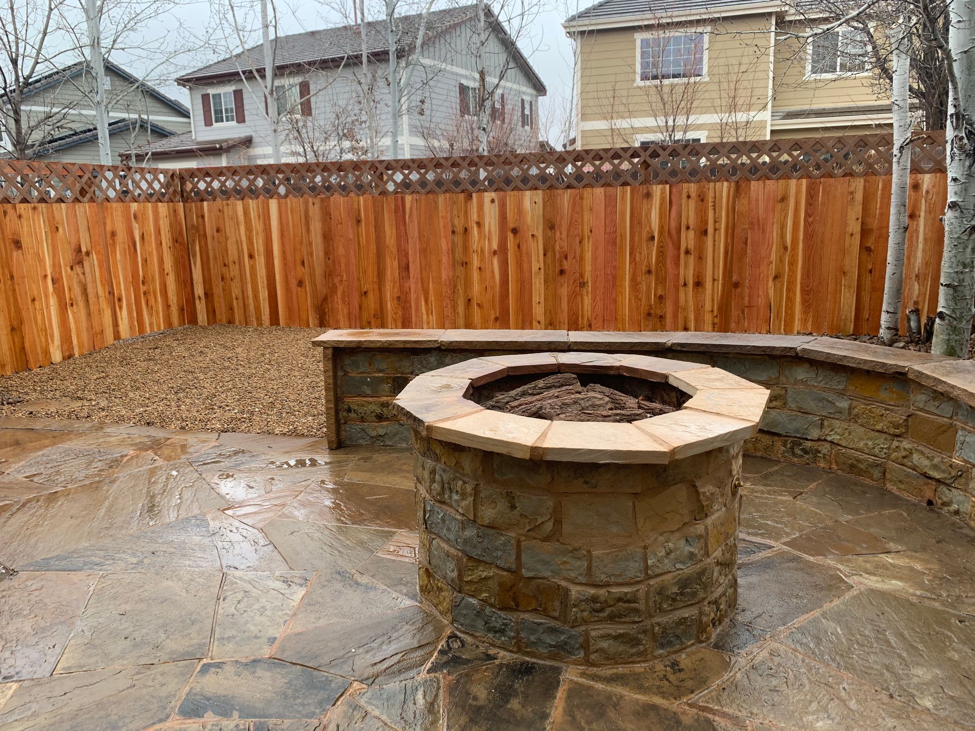 Stone fire pit on a wet patio, surrounded by a low wall and a wooden fence.
