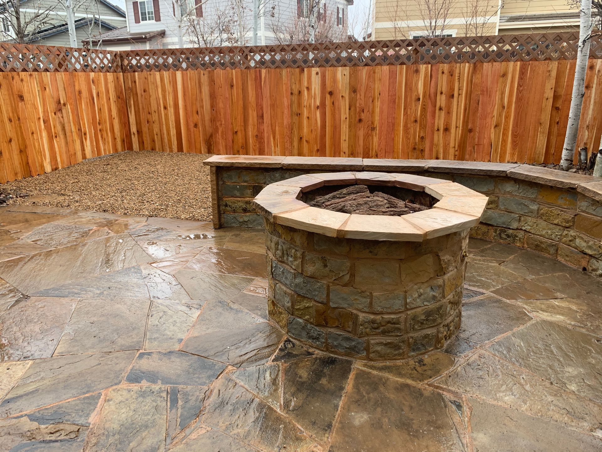 A backyard patio with a stone fire pit, seating, and wooden fence.