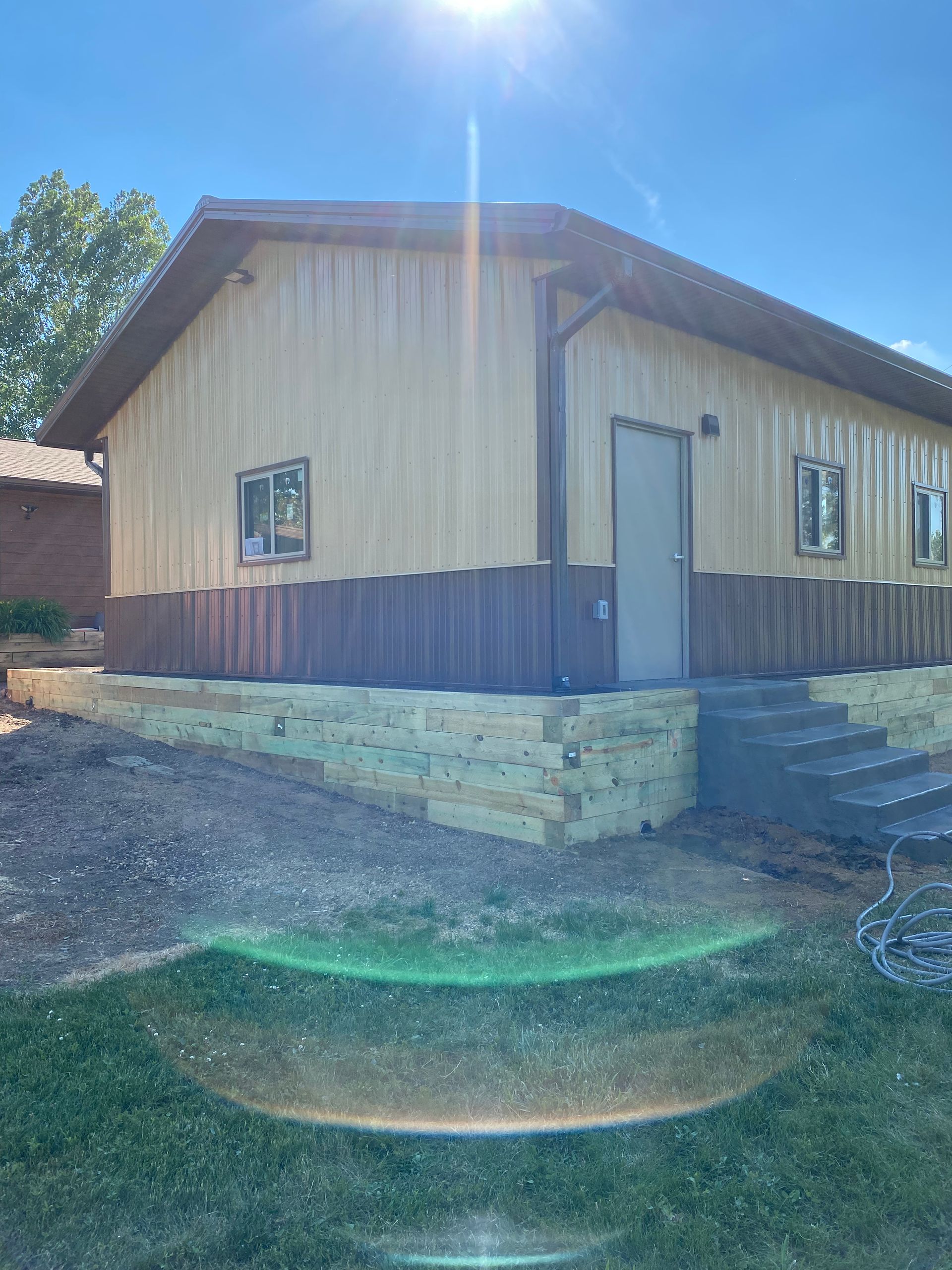 Tan metal-sided building with brown trim, door, and stone base. Two windows. Sunny day.