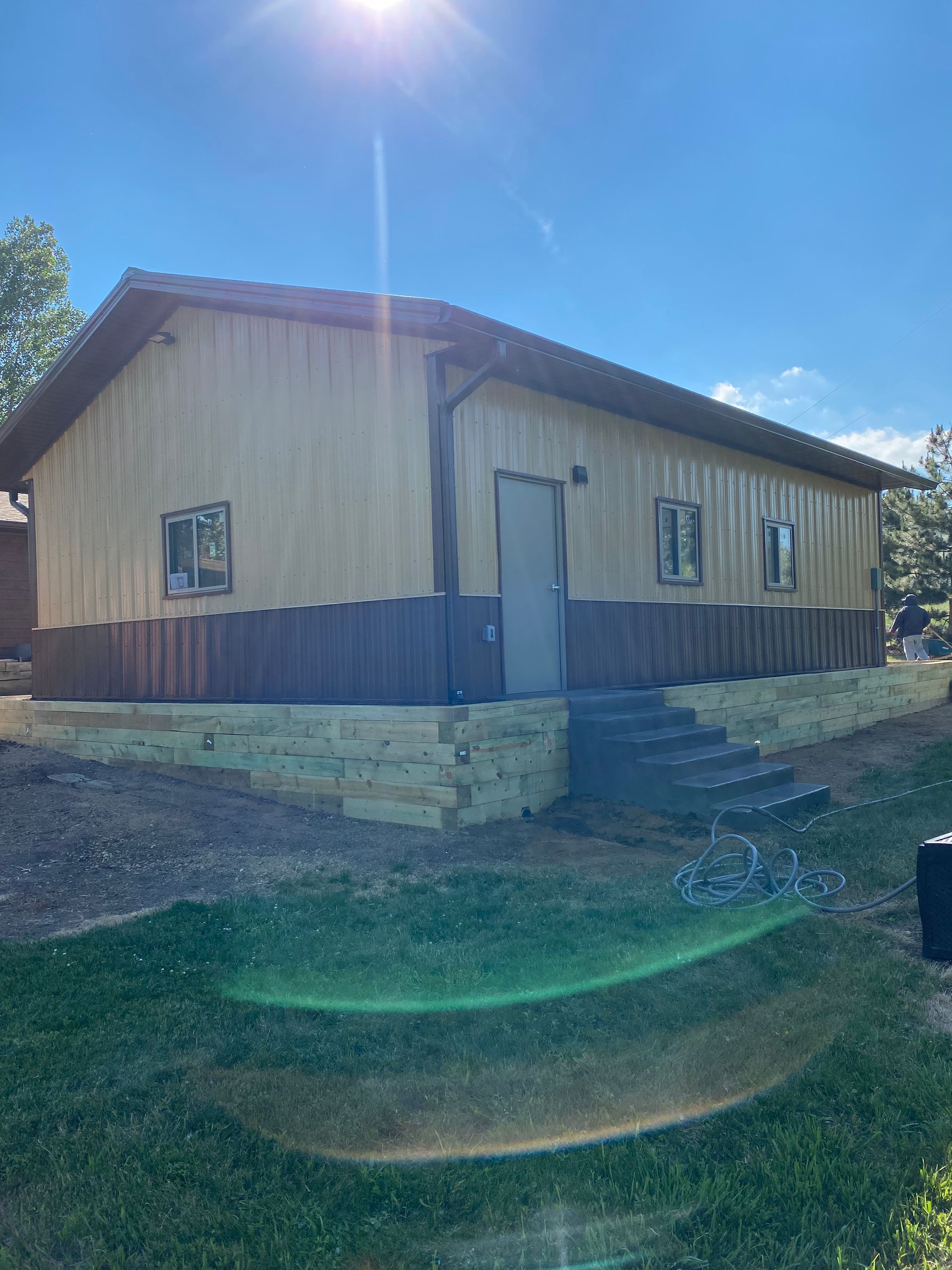 Tan and brown building with a gray door, several windows, and stone retaining wall. Sunny day.