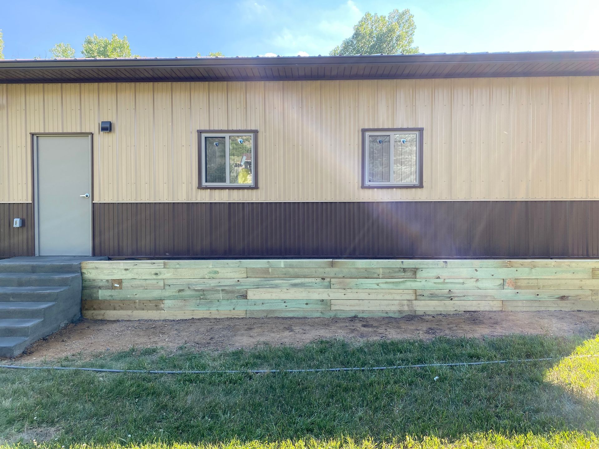 Exterior of a tan and brown building with a wooden retaining wall and grassy lawn. A door and windows are visible.