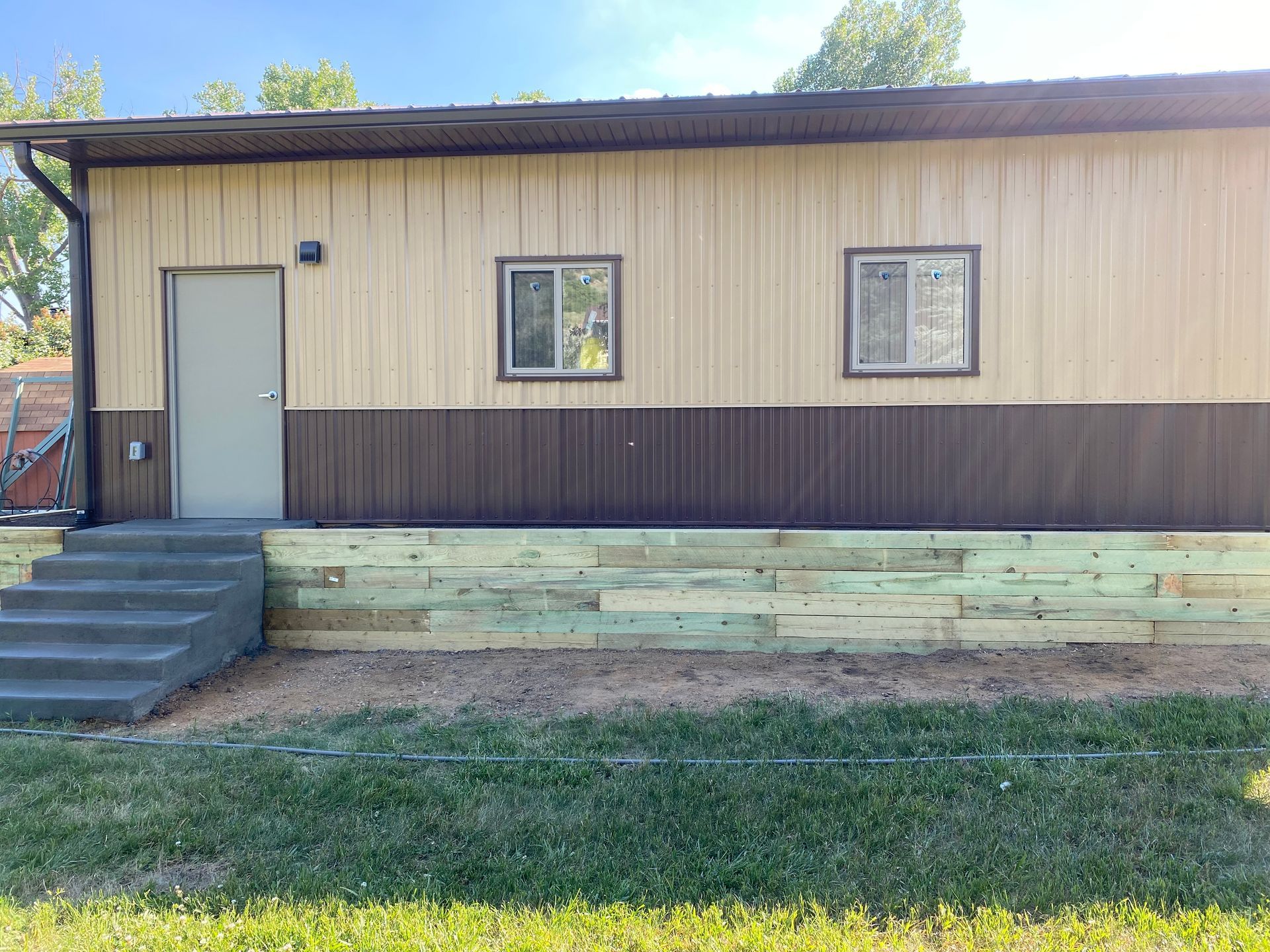 Tan and brown building with steps, door, windows, and a wooden retaining wall.