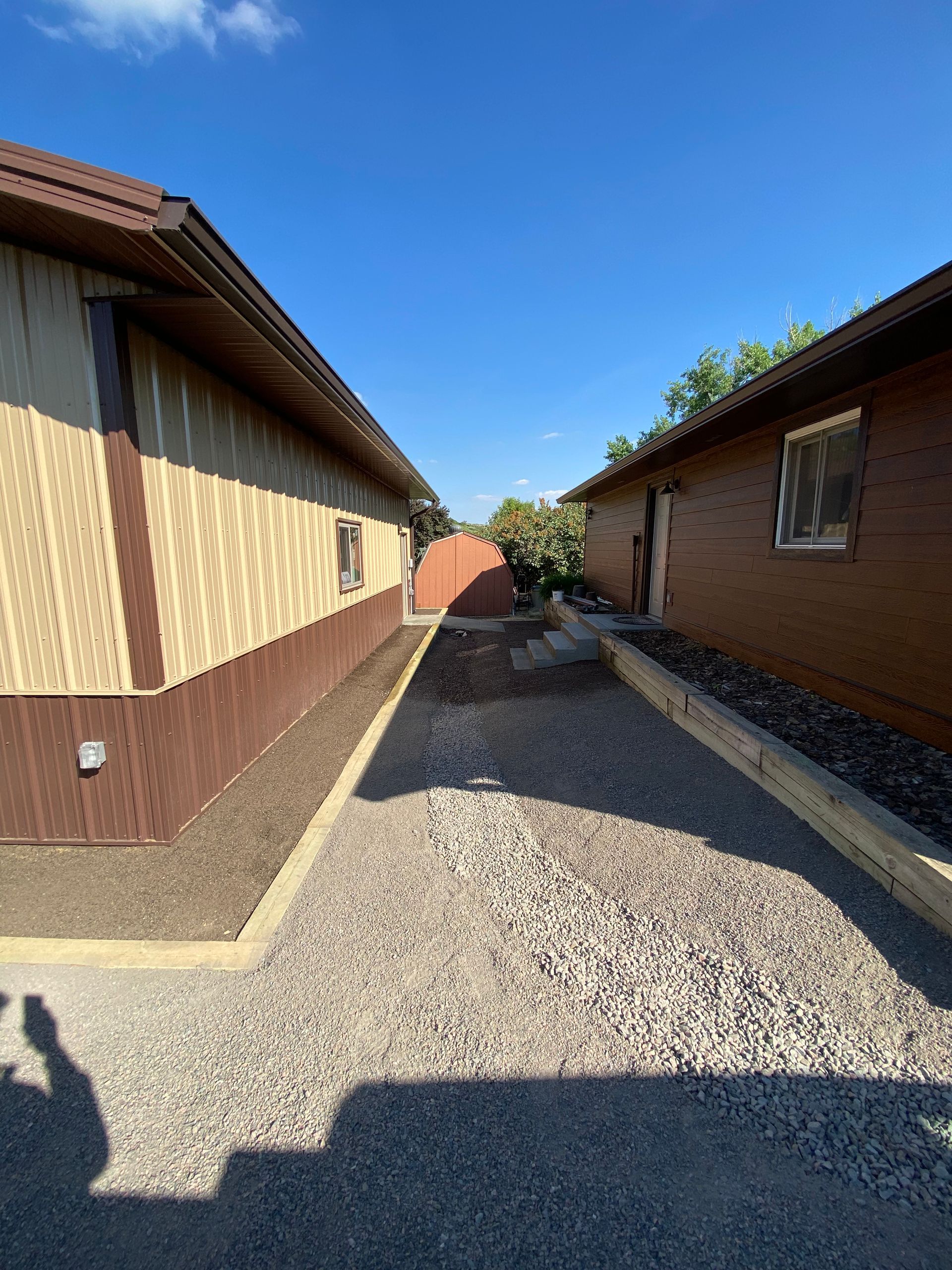 Gravel walkway between two buildings, one with tan siding, the other brown, under a blue sky.