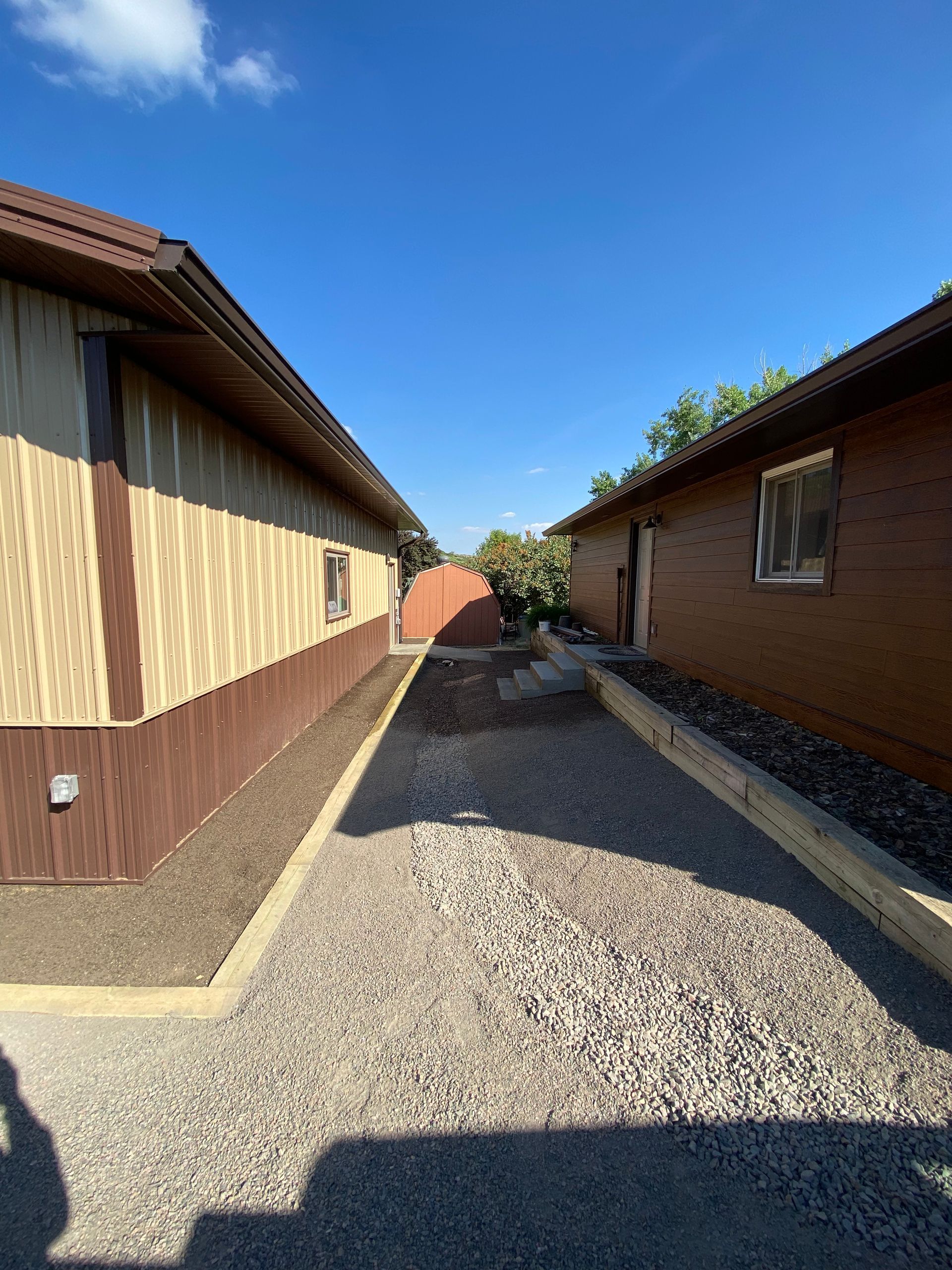 Gravel pathway between two buildings; beige and brown siding. Clear blue sky overhead.