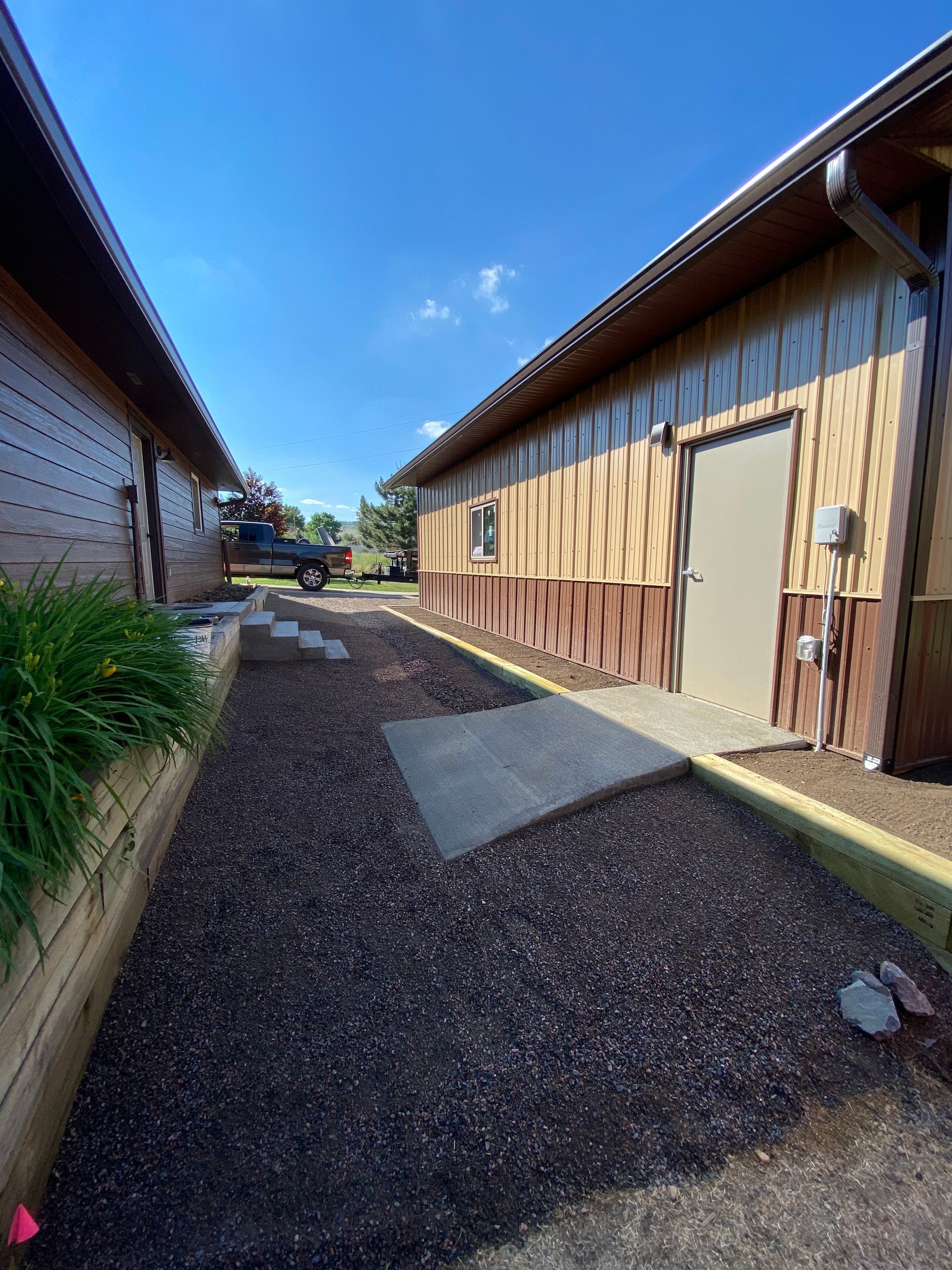 Pathway between two buildings with brown siding, gravel ground, and blue sky.