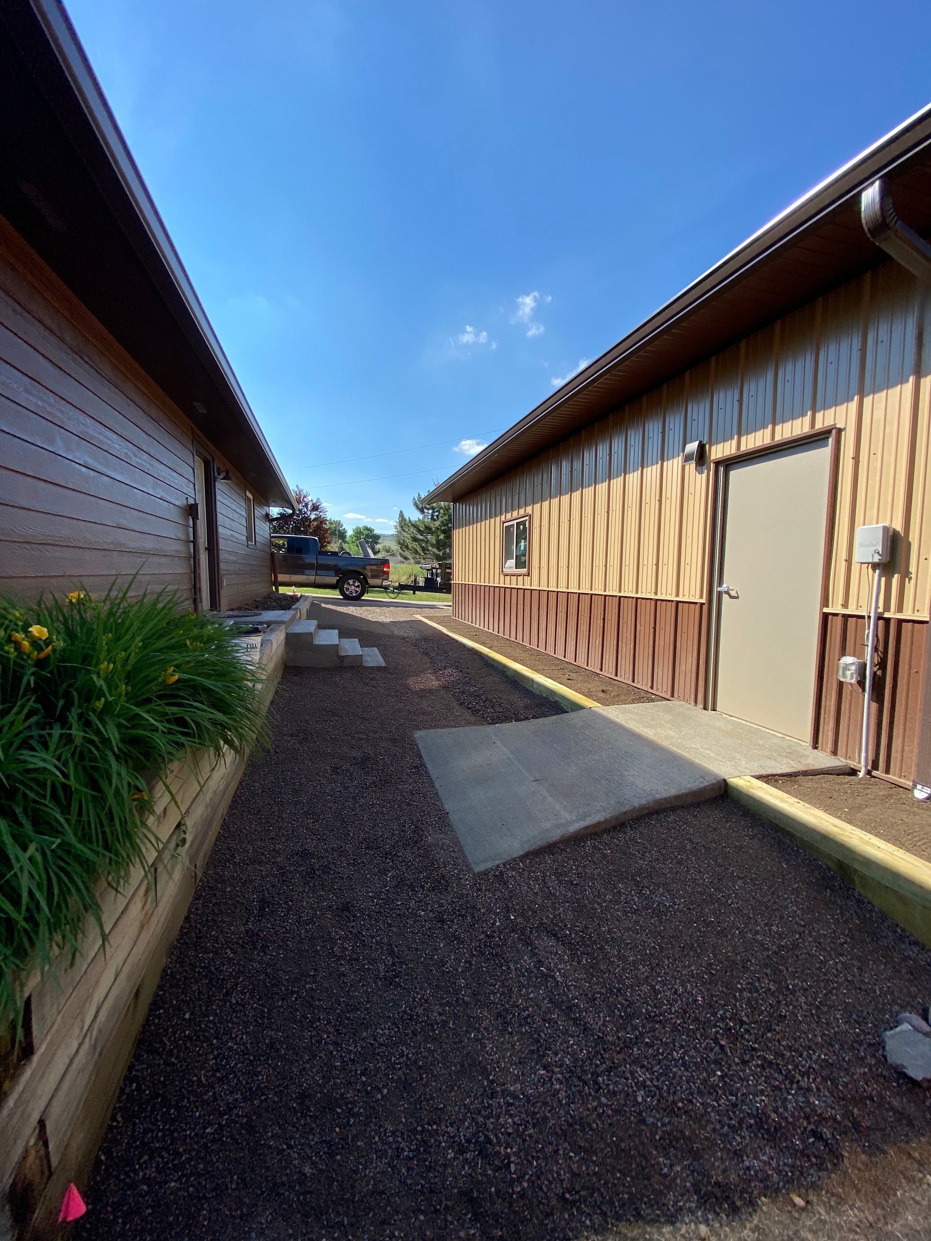 Pathway between two buildings with mulch ground cover, small concrete sections, and a truck in the distance under a blue sky.