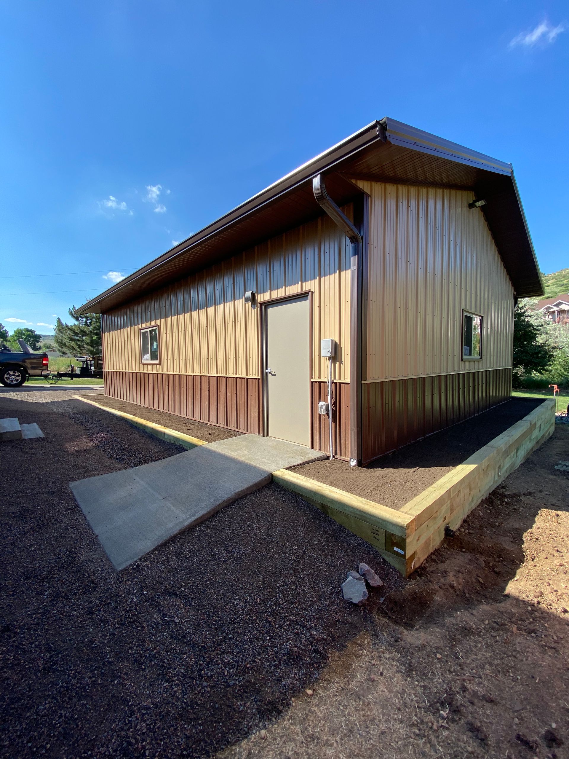 Tan and brown metal building with a concrete walkway, on a gravel bed, under a blue sky.