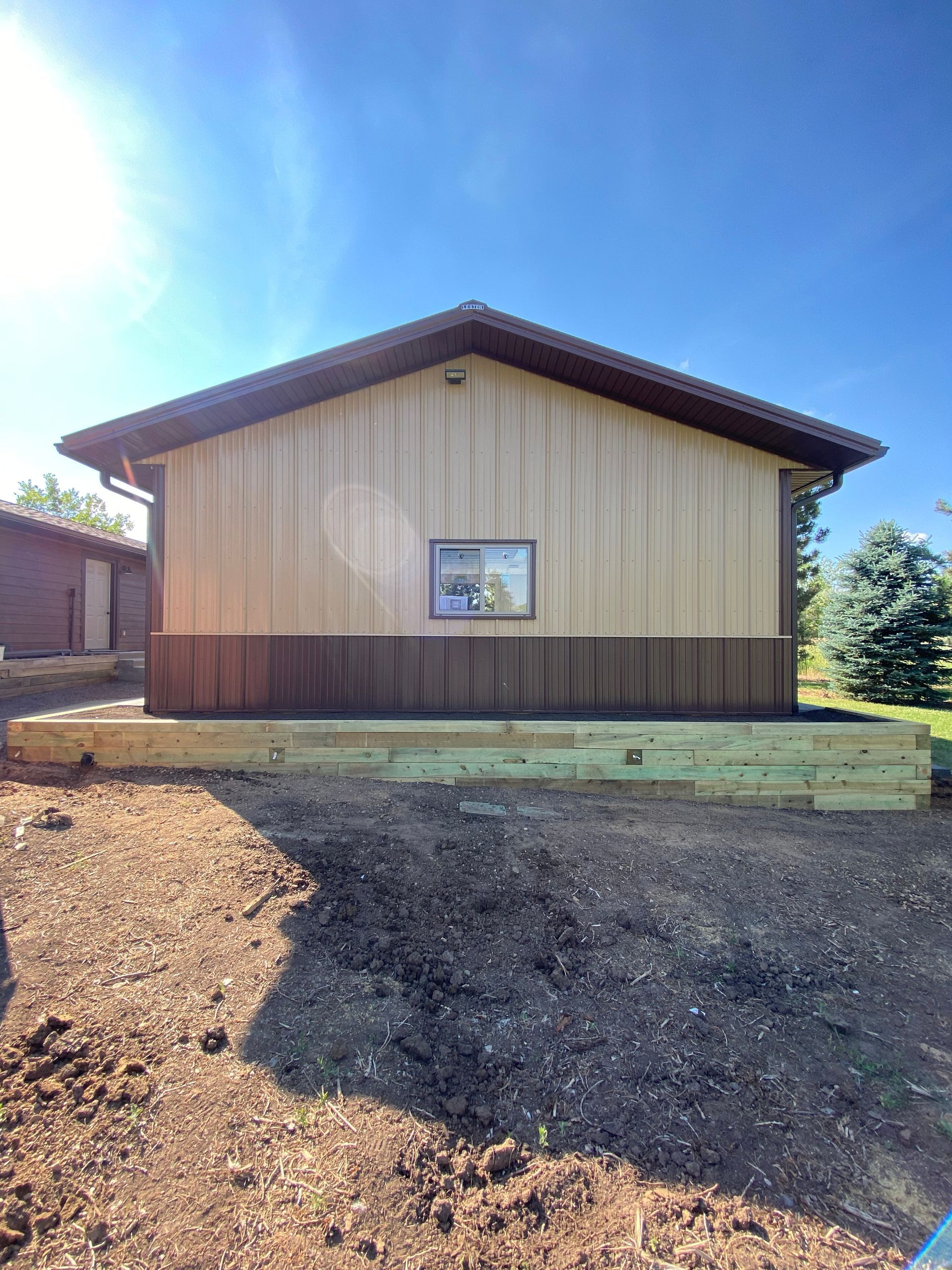 Tan and brown building with a single window. Wooden retaining wall in front, dirt and wood chips below.