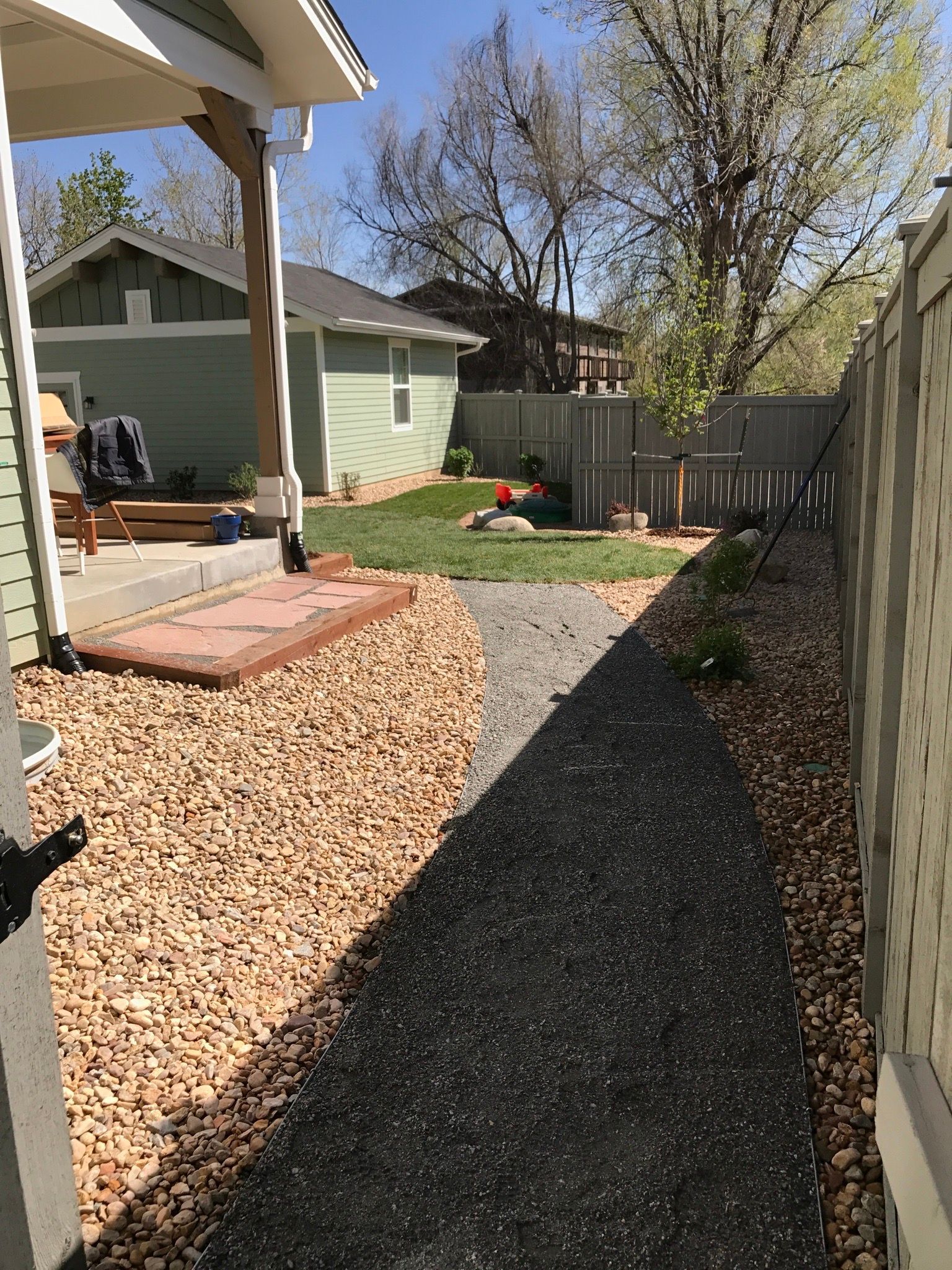 A narrow gravel path leads to a grassy area, flanked by beige gravel and a wooden fence. Sunny outdoor scene.