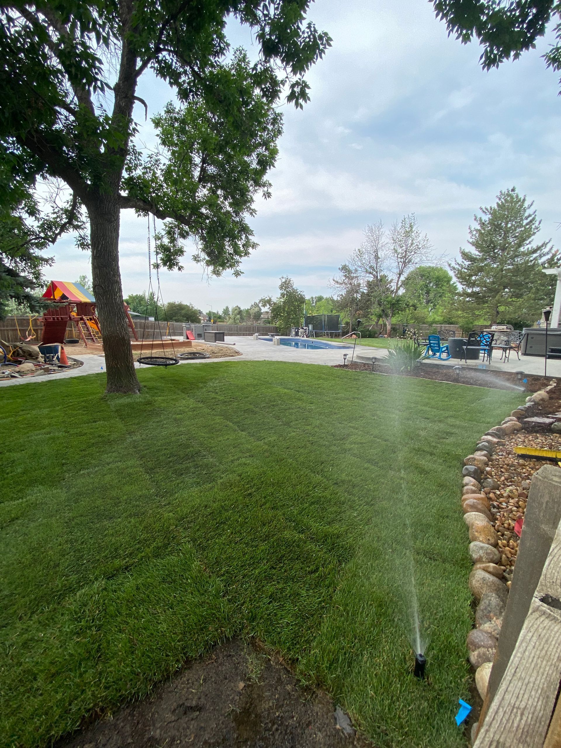 Green lawn being watered; backyard with a pool, trees, and cloudy sky.