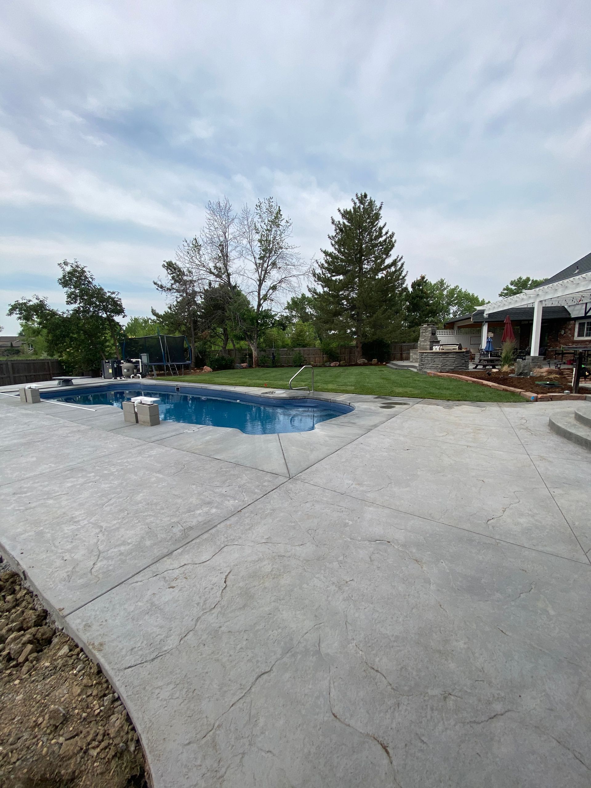 Concrete patio next to a blue swimming pool, with a house and trees in the background. Overcast sky.