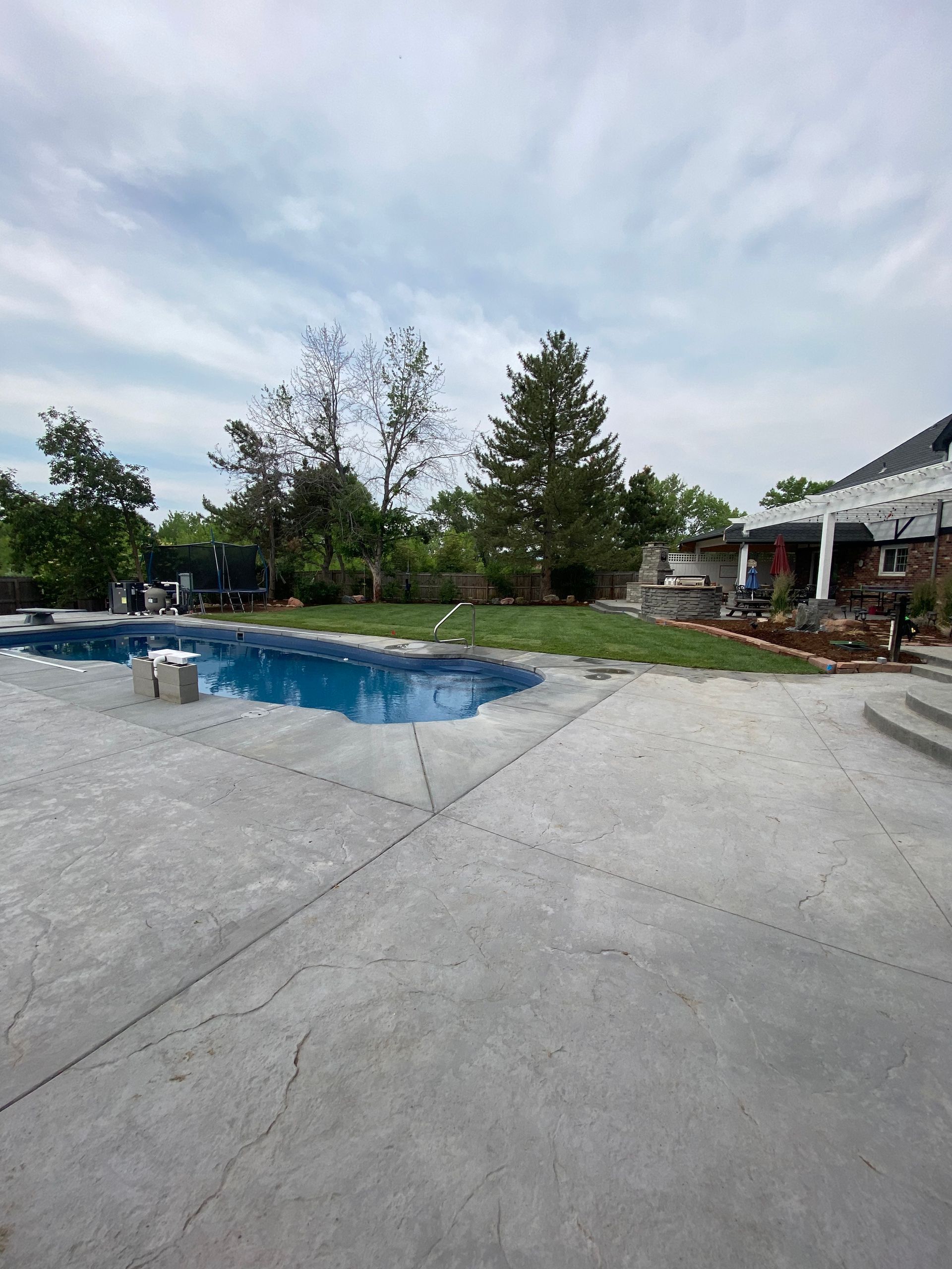 Concrete patio with a pool on the left and a yard with trees in the back under a cloudy sky.