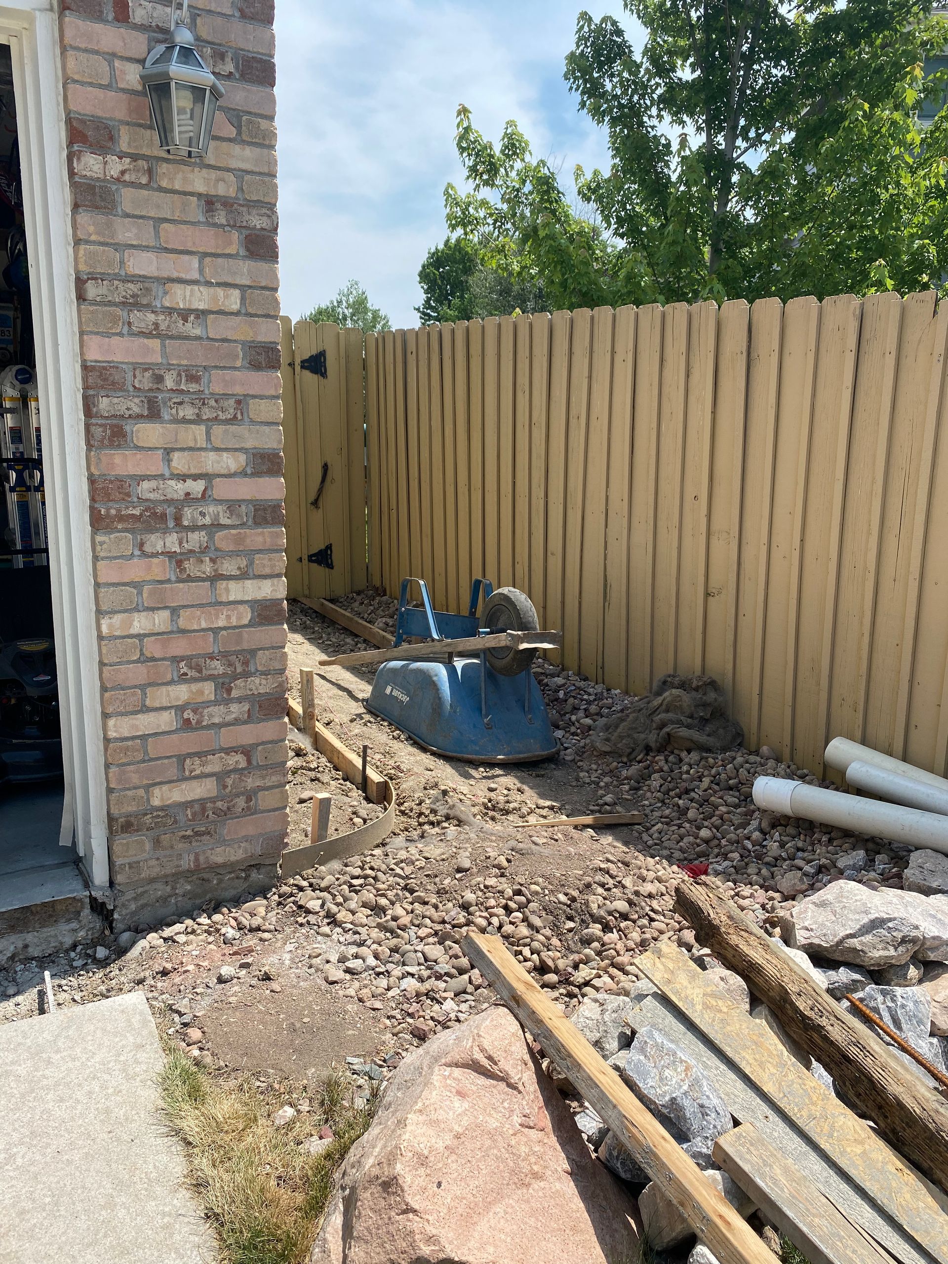 Garage, fence, and construction materials in a yard. Blue wheelbarrow on gravel pathway, wooden fence, brick garage.