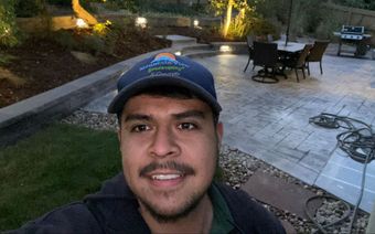 Man smiling, selfie in a backyard with patio, lit by outdoor lights at dusk.