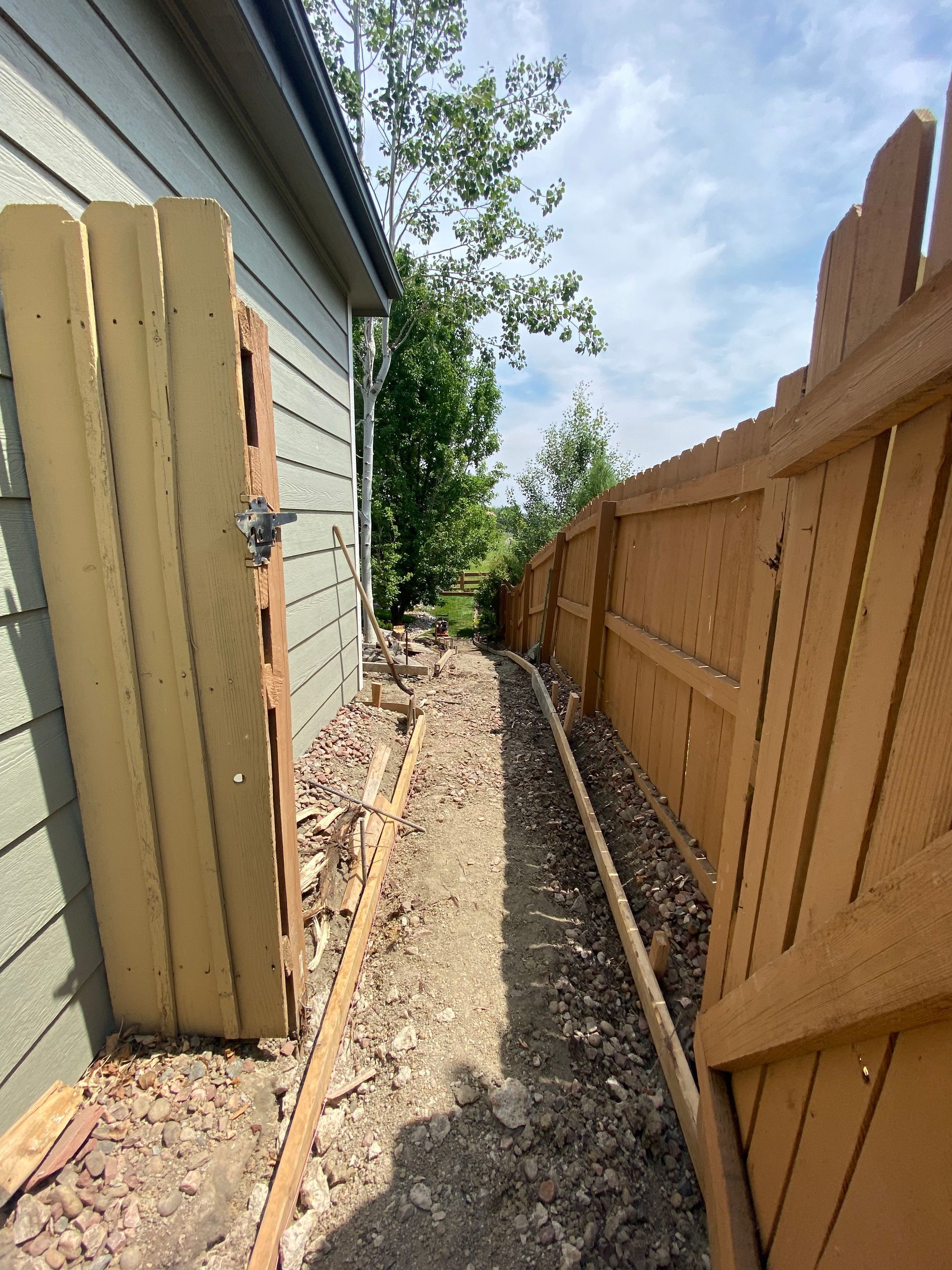 A narrow dirt path between two wooden fences and a light green building on a sunny day.