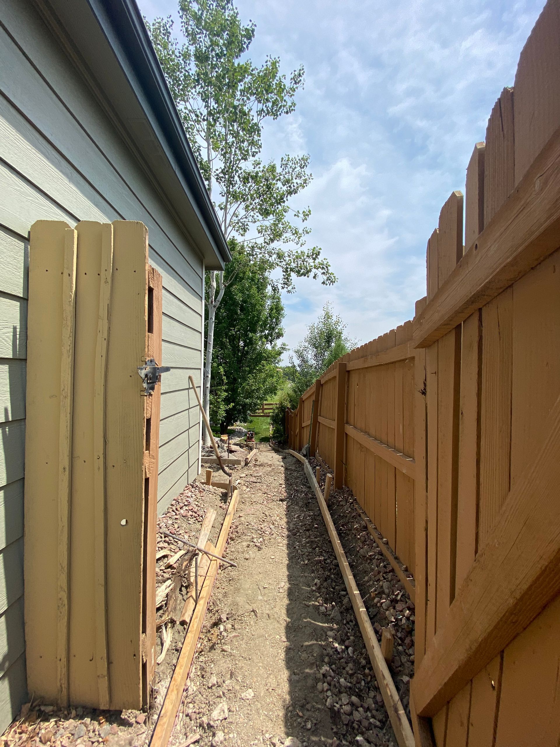 Narrow, dirt pathway between a house (left) and wooden fence (right), leading to a bright, sunny sky.