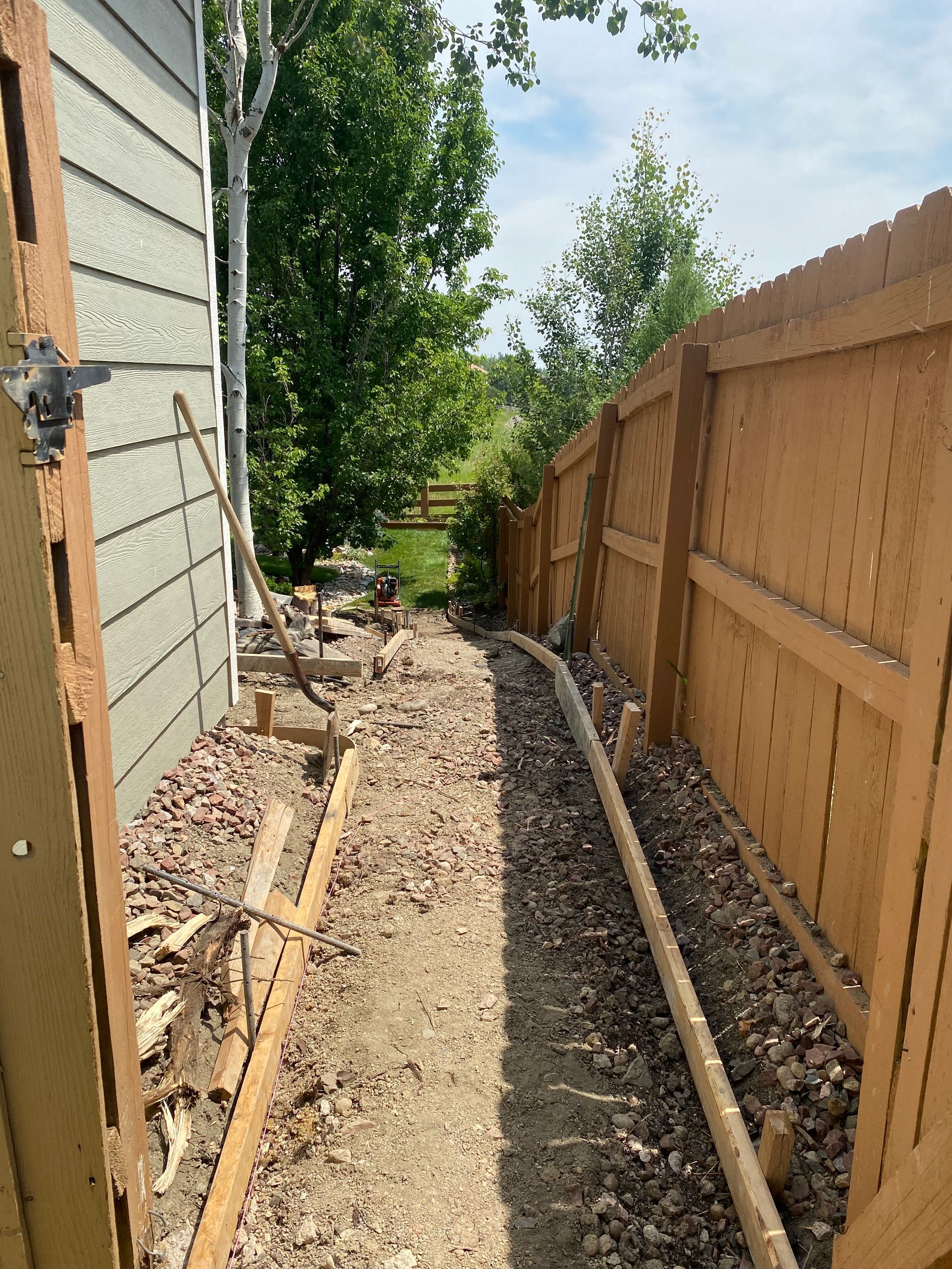 Narrow dirt path between a wooden fence and a house, with concrete forms and landscaping materials.