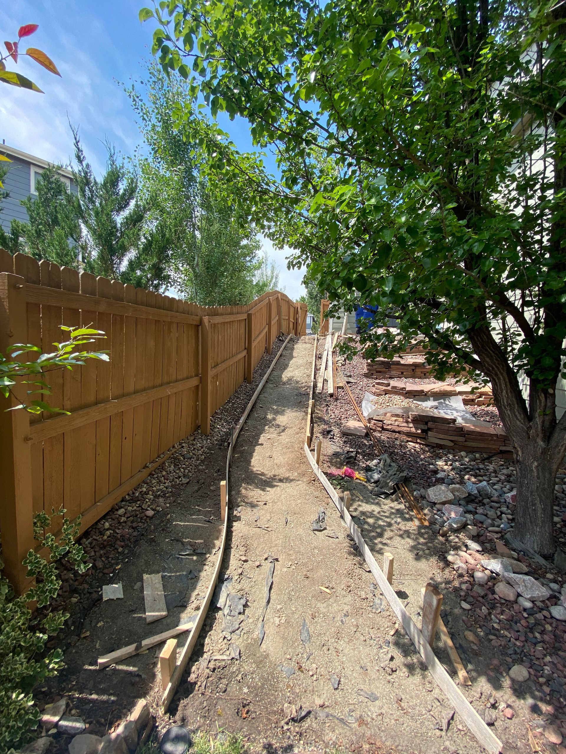Narrow dirt path between a wooden fence and a partially demolished area with a tree and blue sky.