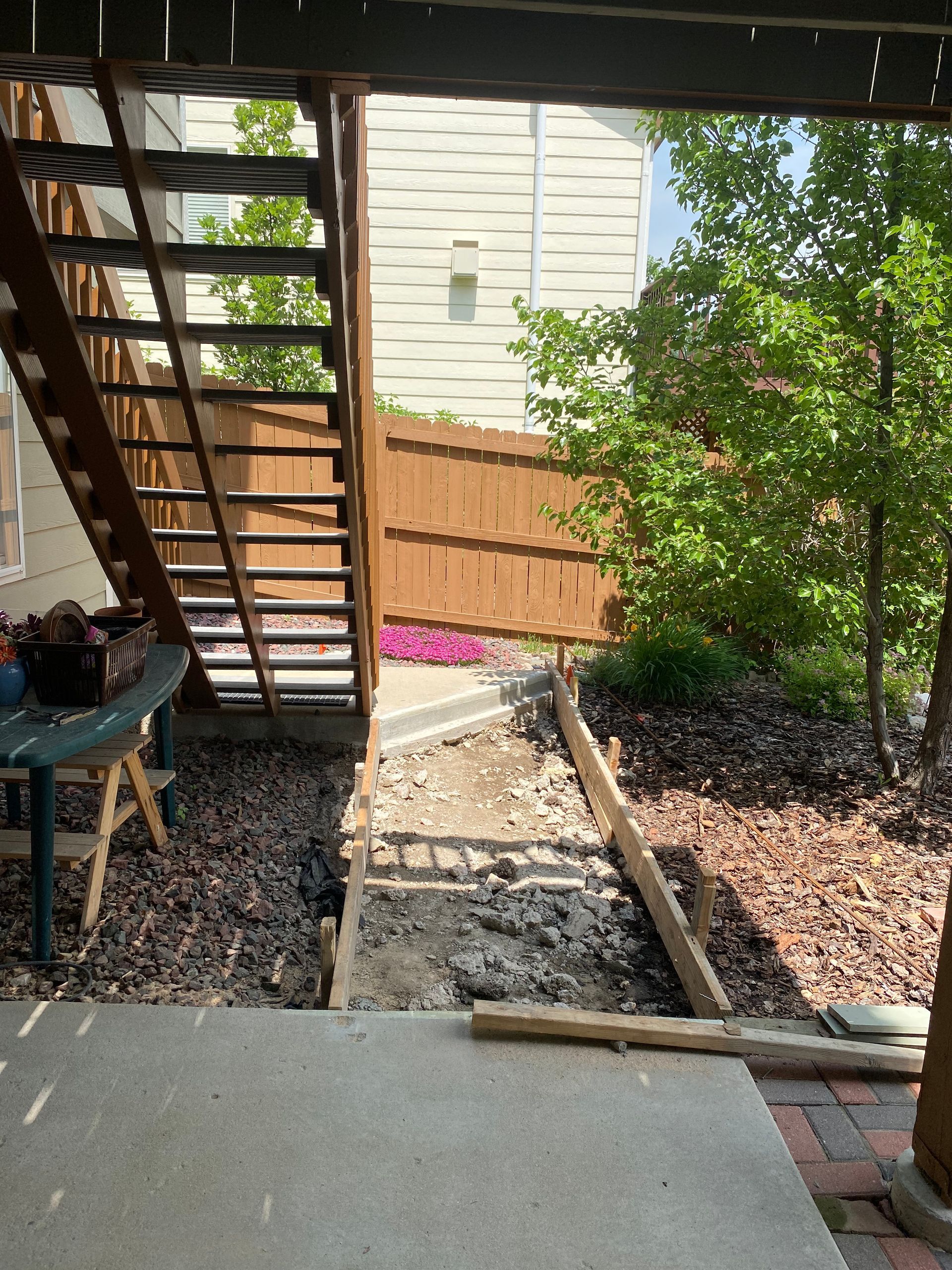 Pathway under stairs with exposed dirt, leading to a fence and trees. A deck overhangs the area.